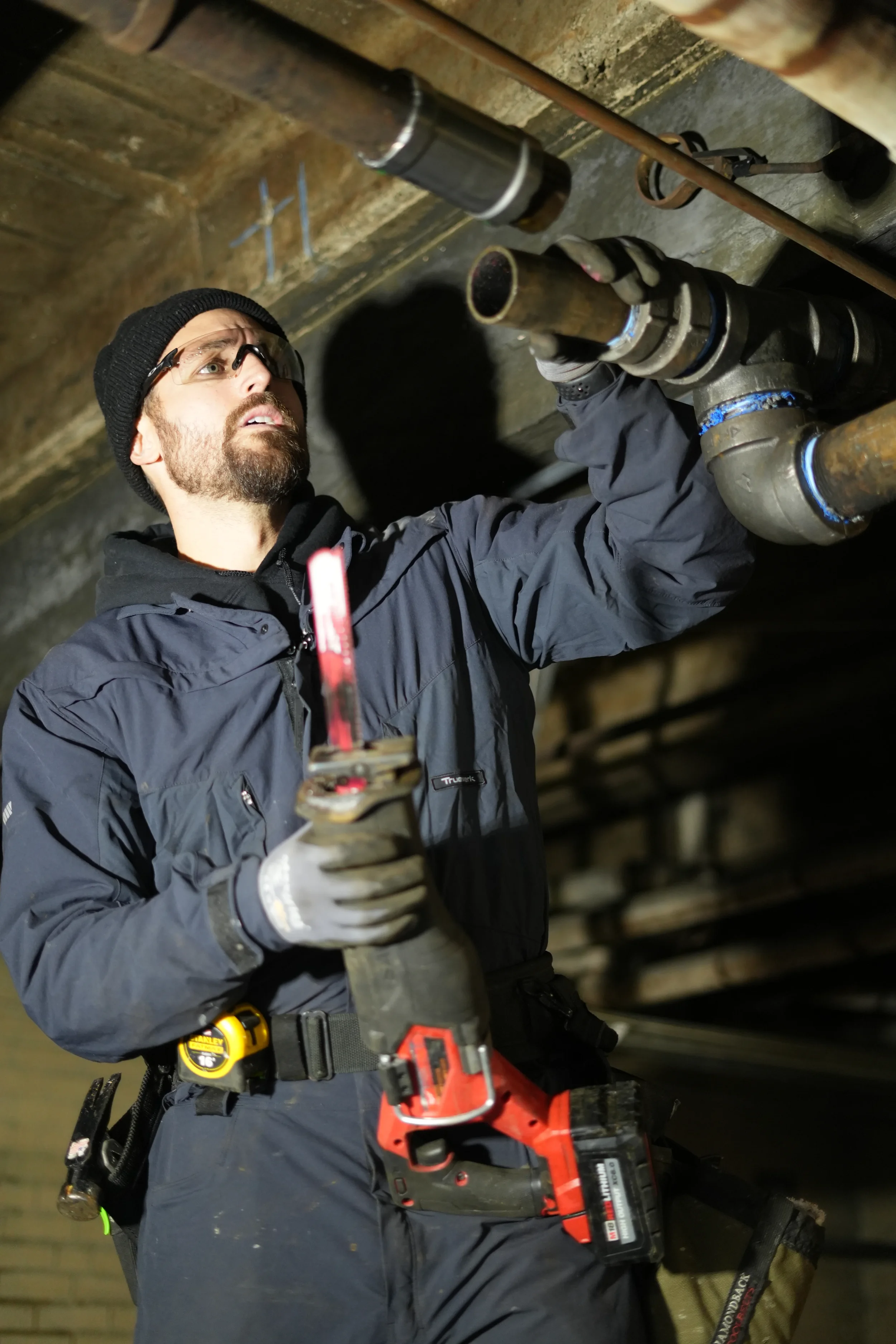 A man wearing safety glasses working on plumbing pipes underground, holding a cordless power drill.