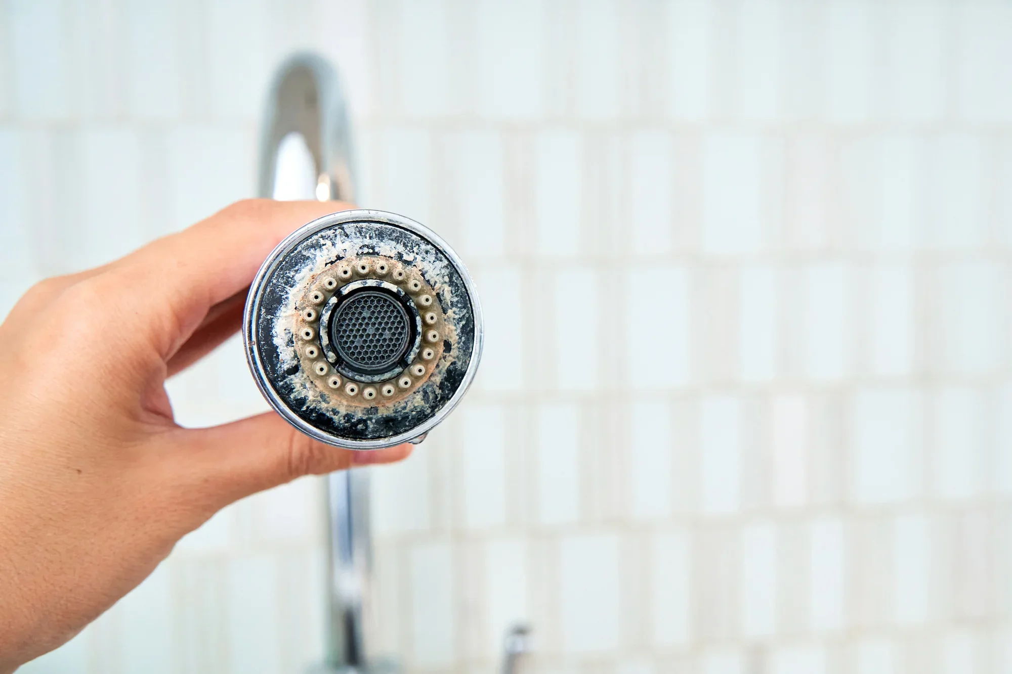 Close-up view of a person's hand holding a faucet aerator in a kitchen or bathroom sink with a white tiled wall in the background.