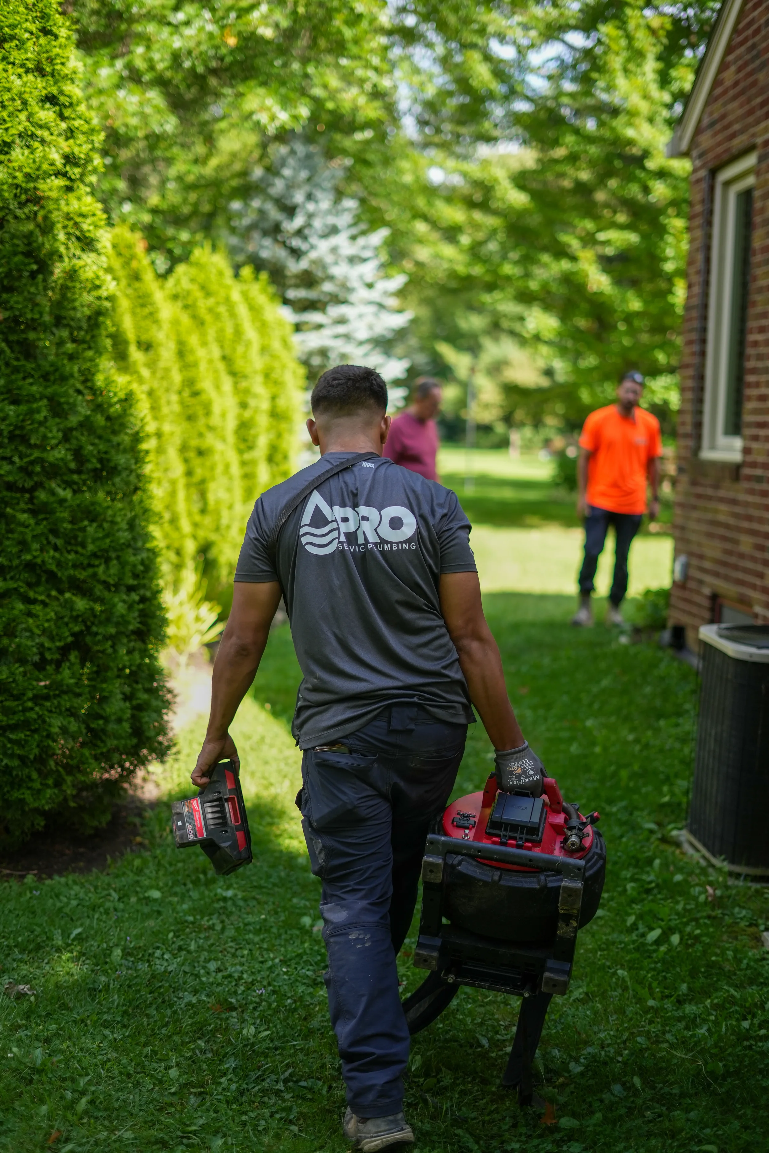 A worker carrying a piece of equipment in a backyard, with two other people in the background.