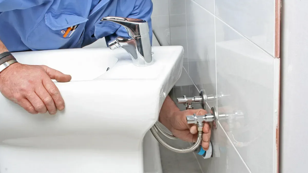 A person installing plumbing fixtures behind a bathroom sink, with one hand on the sink and the other on the water valves.