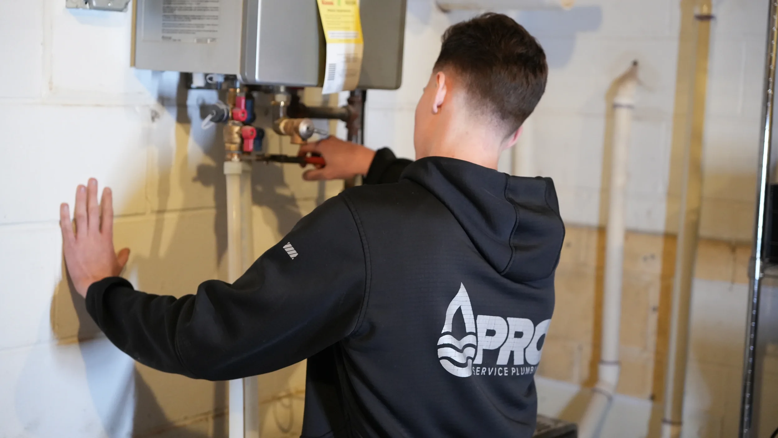 A technician in a black hoodie labeled 'Pro Service Plumbing' repairing plumbing pipes in a utility room.