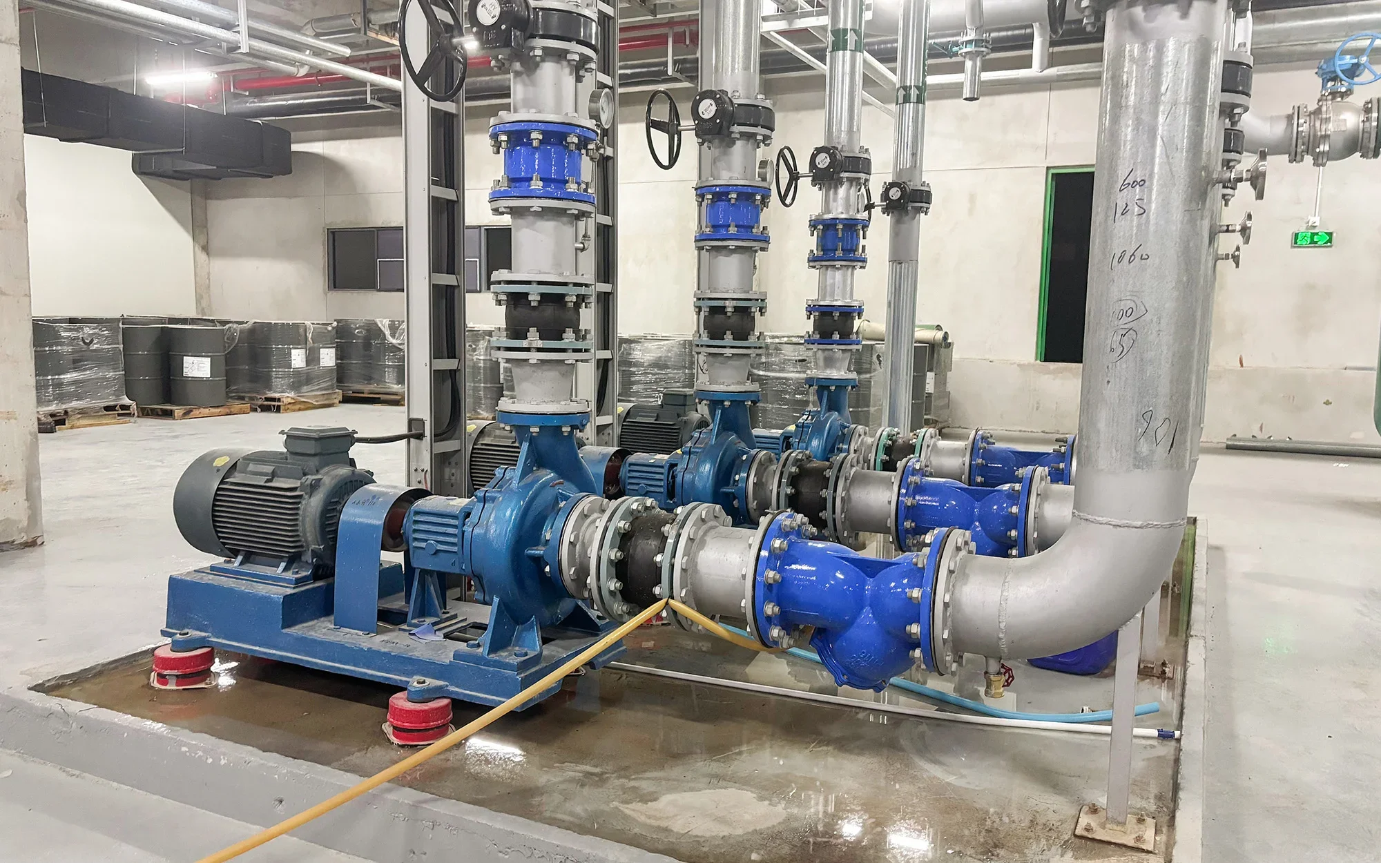 An industrial mechanical setup with large blue and silver pipes, valves, and motors in a clean, well-lit room with concrete floors and walls. The pipes are marked with handwritten measurements and the setup is secured on a raised concrete platform.