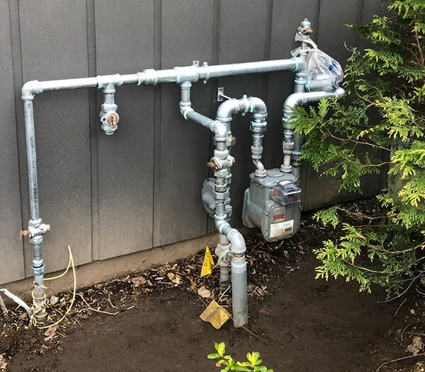 Outdoor natural gas meter and pipes installed against a house wall, with surrounding soil and some green shrubs.