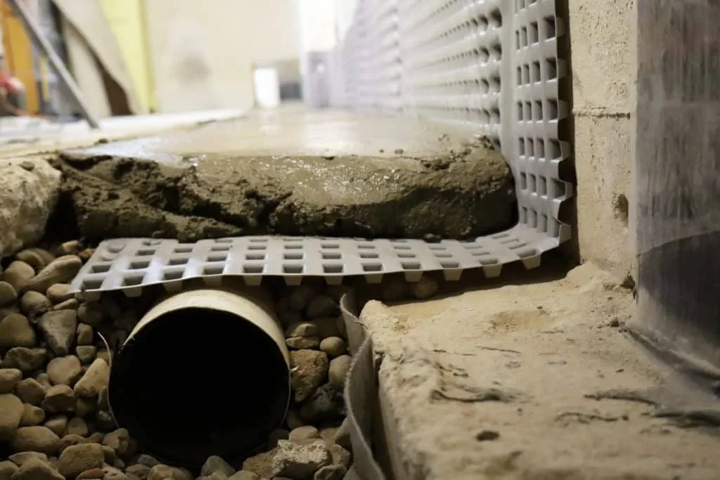 Close-up of a drain pipe installed under a concrete foundation, with gravel and a drainage grate nearby.
