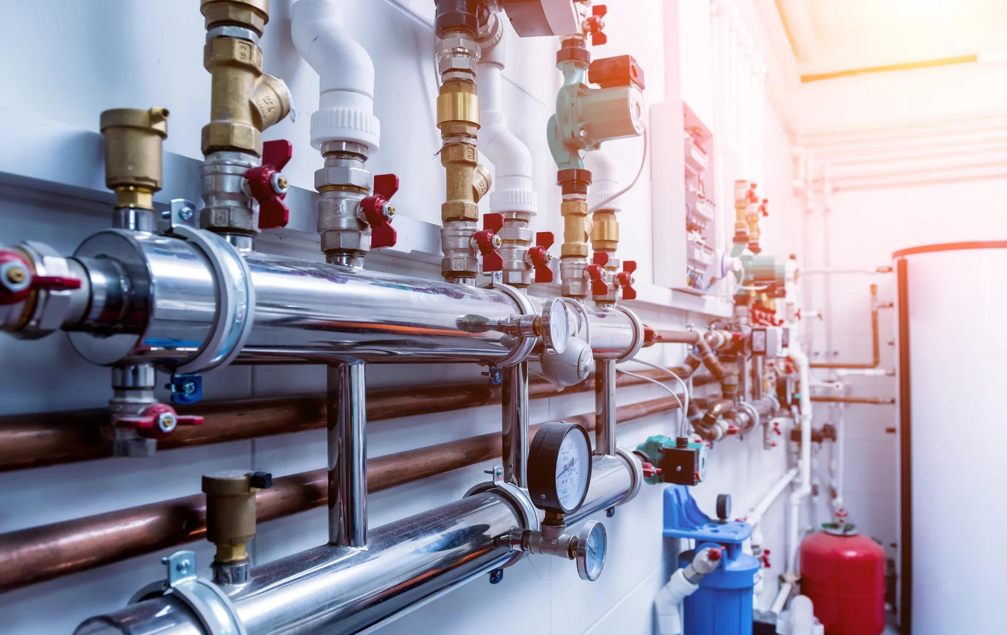 Plumbing and piping system with valves, gauges, and pipes in a utility room, illuminated by natural light.