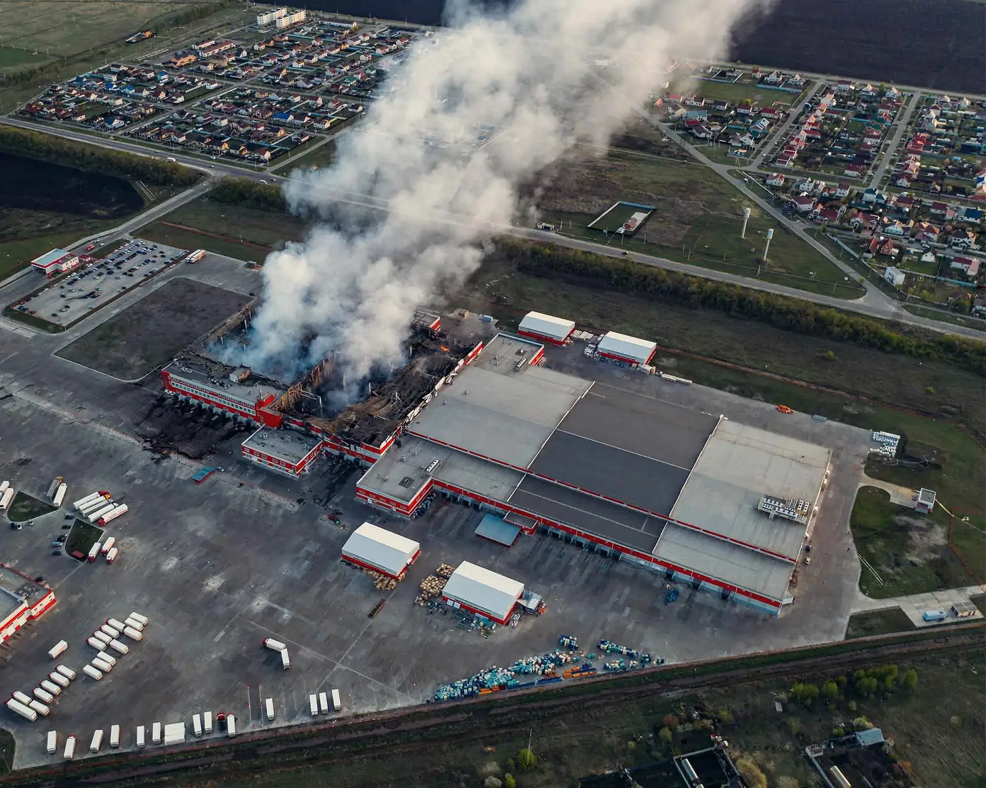 An aerial view of a large building fire with thick smoke rising from a section of the structure, surrounded by parking lots and residential neighborhoods.