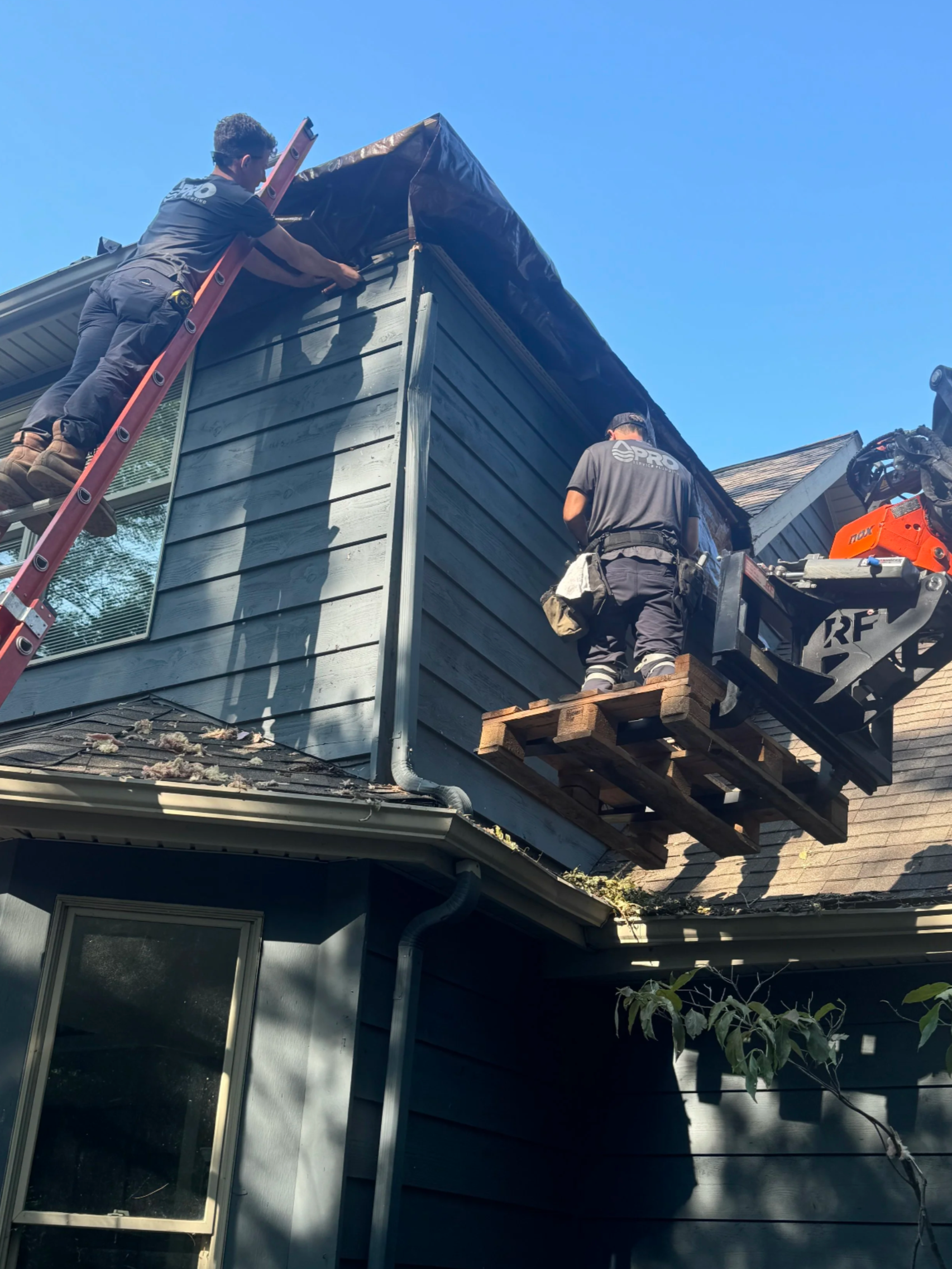 Two workers are repairing the exterior of a two-story house, using a lift and a ladder on a sunny day.