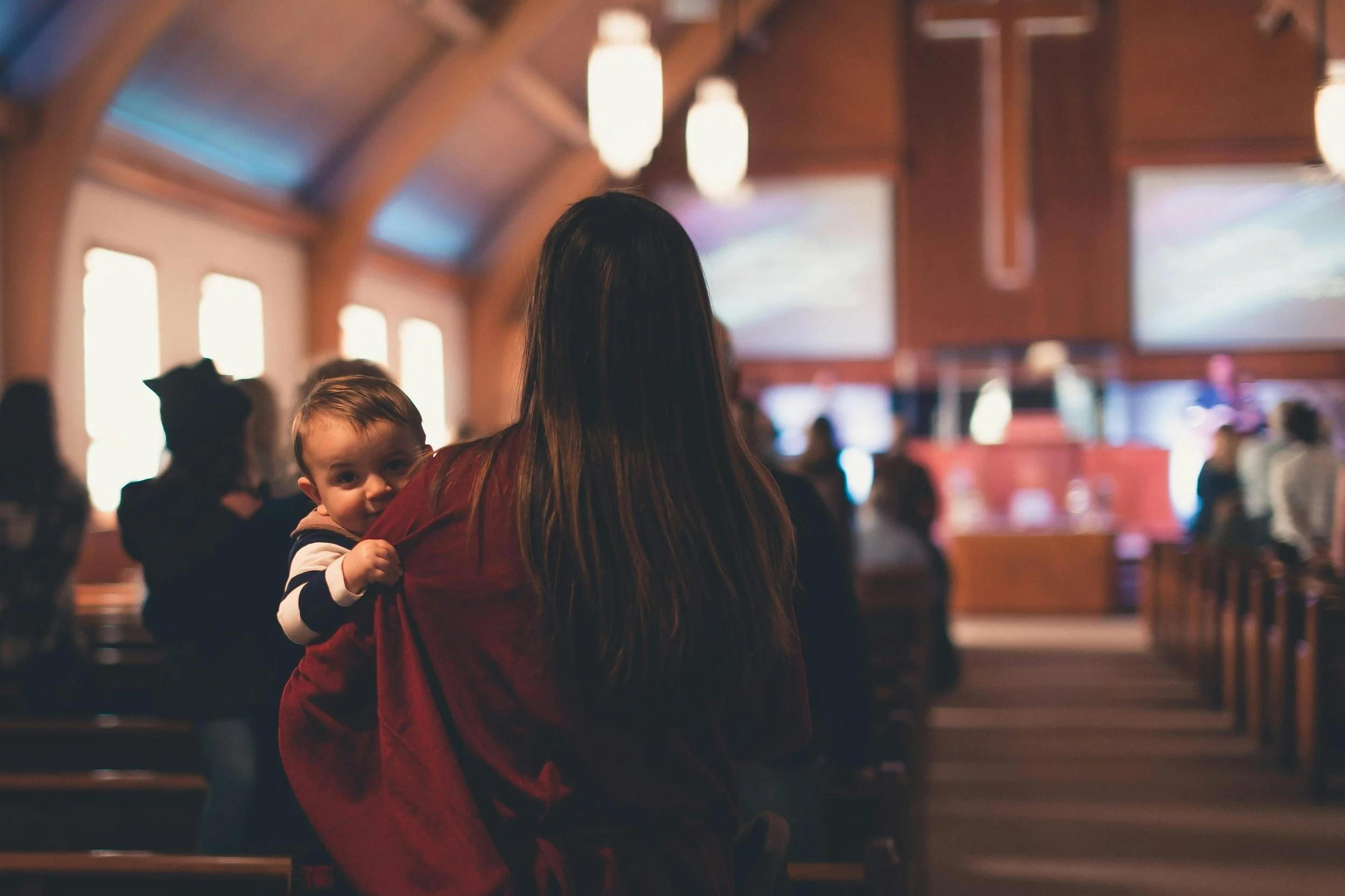 A woman with long hair holding a small child inside a church or chapel, with blurred figures and wooden interior in the background.
