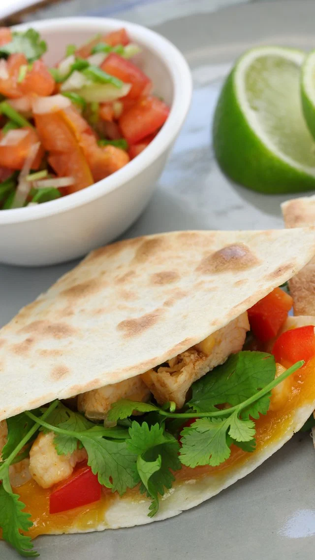 A taco with grilled chicken, red bell peppers, cilantro, and orange sauce, accompanied by a lime wedge, and a bowl of pico de gallo with chopped tomatoes, onions, and green herbs.