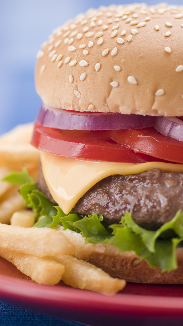 Close-up of a cheeseburger with sesame seed bun, lettuce, tomato, cheese, beef patty, and red onion slices, with French fries on the side.