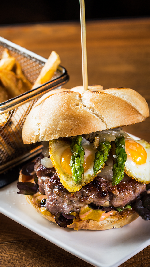 Close-up of a gourmet burger with two sunny-side-up eggs, green asparagus spears, a beef patty, and lettuce on a toasted bun, served on a white plate with a side of French fries in a basket.