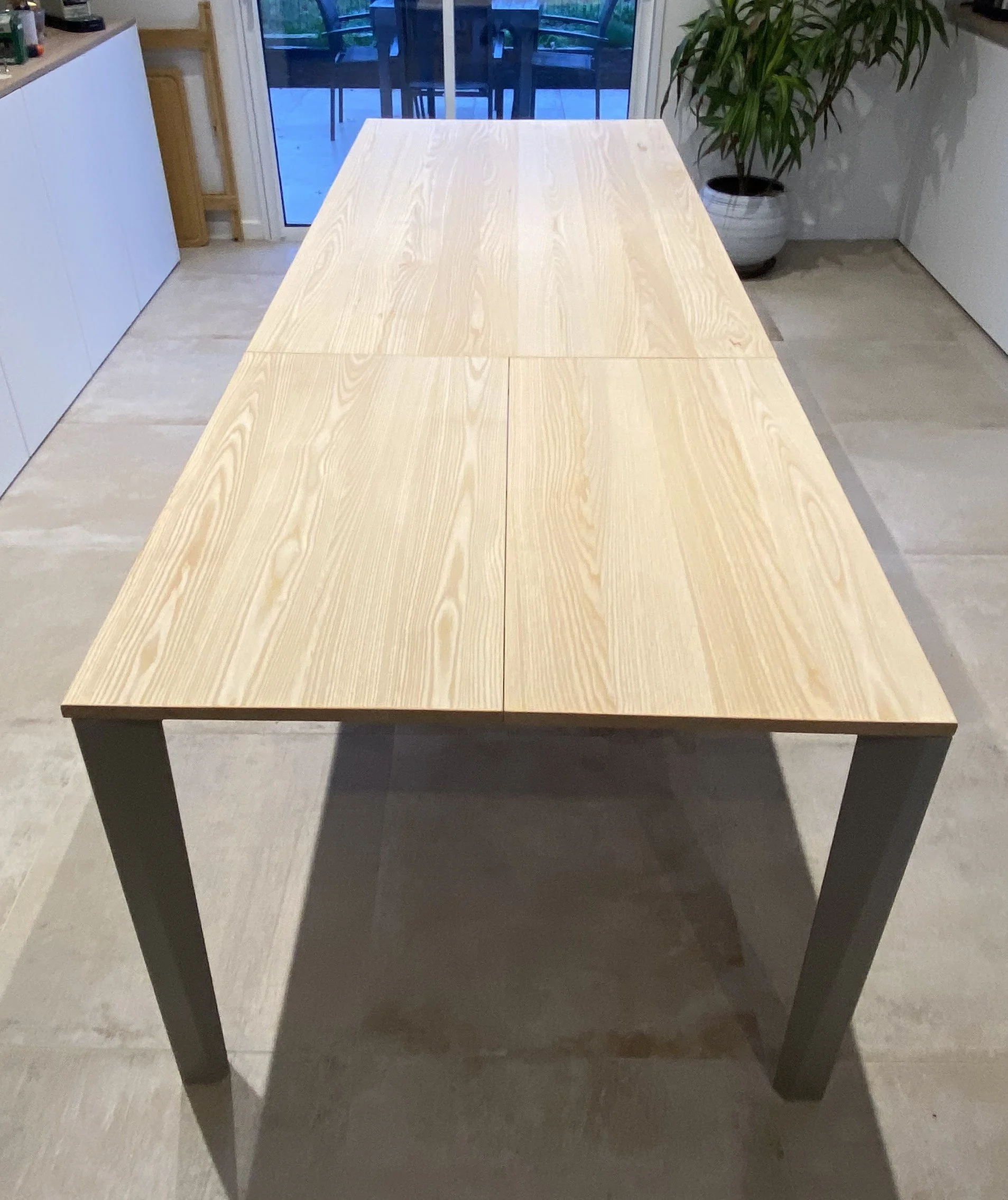 A wooden dining table in a modern kitchen with a sliding glass door leading outside, a potted plant nearby, and tiled flooring.