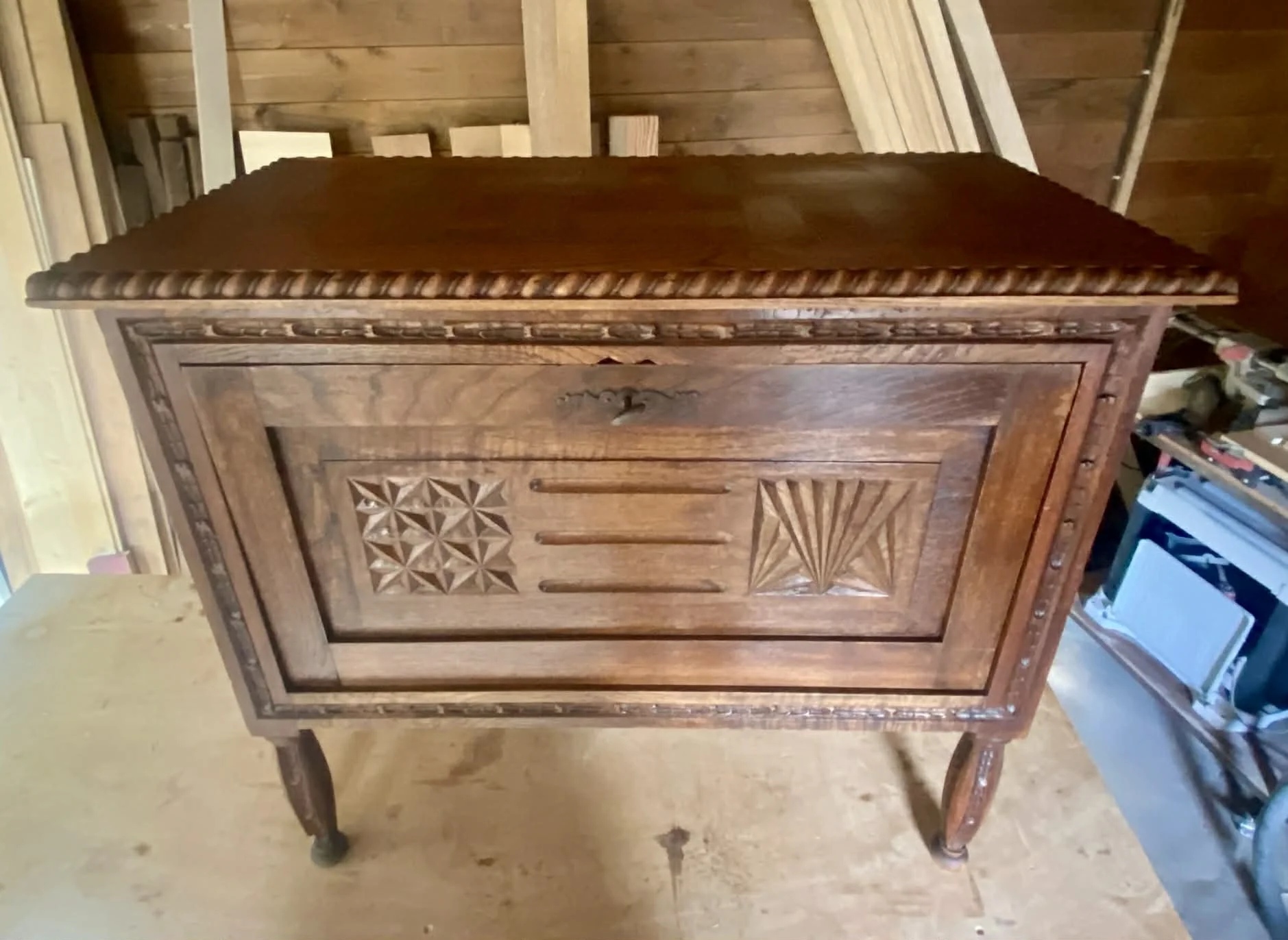 A wooden cabinet with carved geometric patterns on the door, standing on four curved legs in a woodworking workshop.