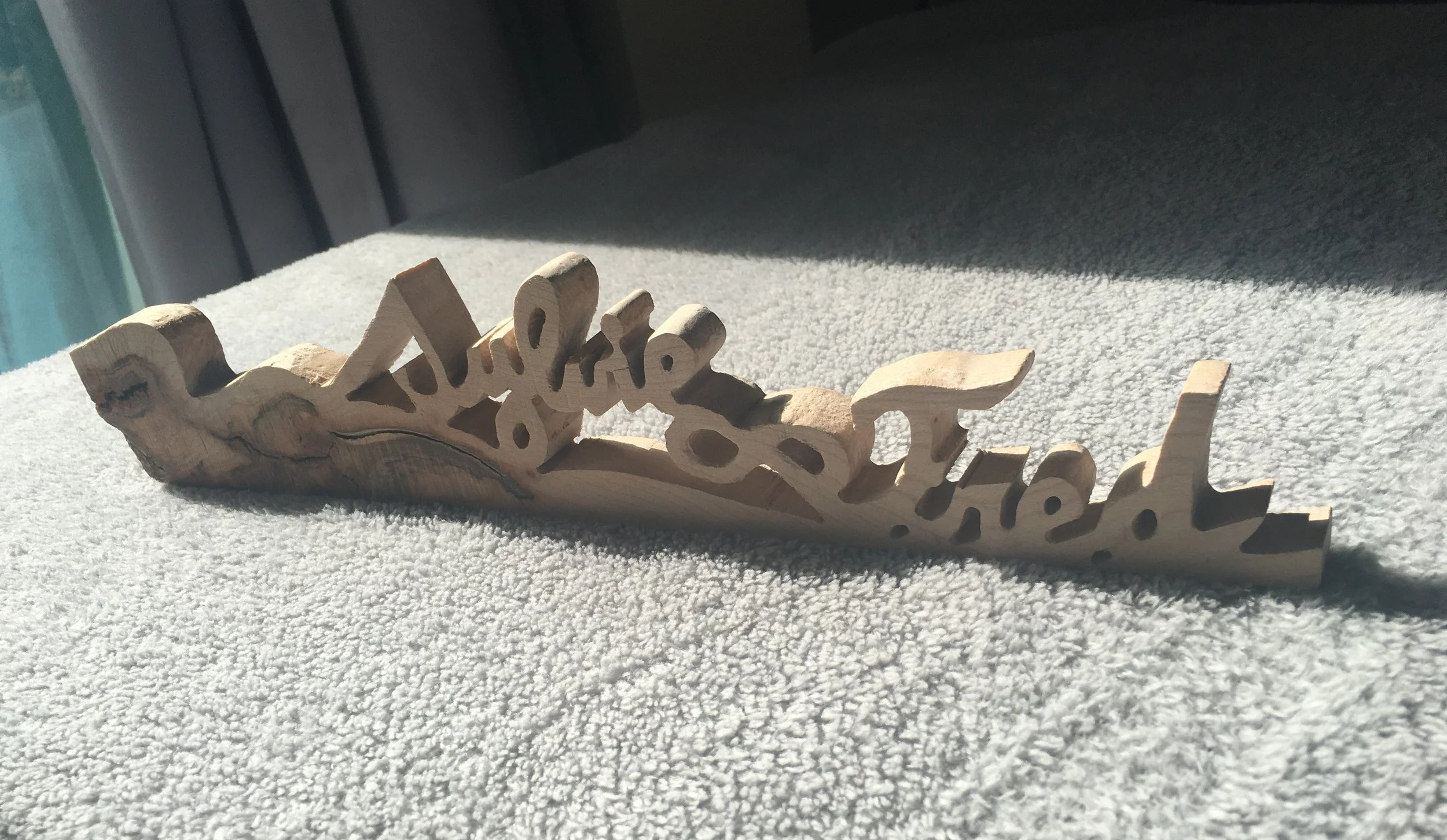 Wooden decorative piece with cutout letters spelling 'Sylvie&Fred' resting on a textured gray fabric surface with sunlight casting shadows.