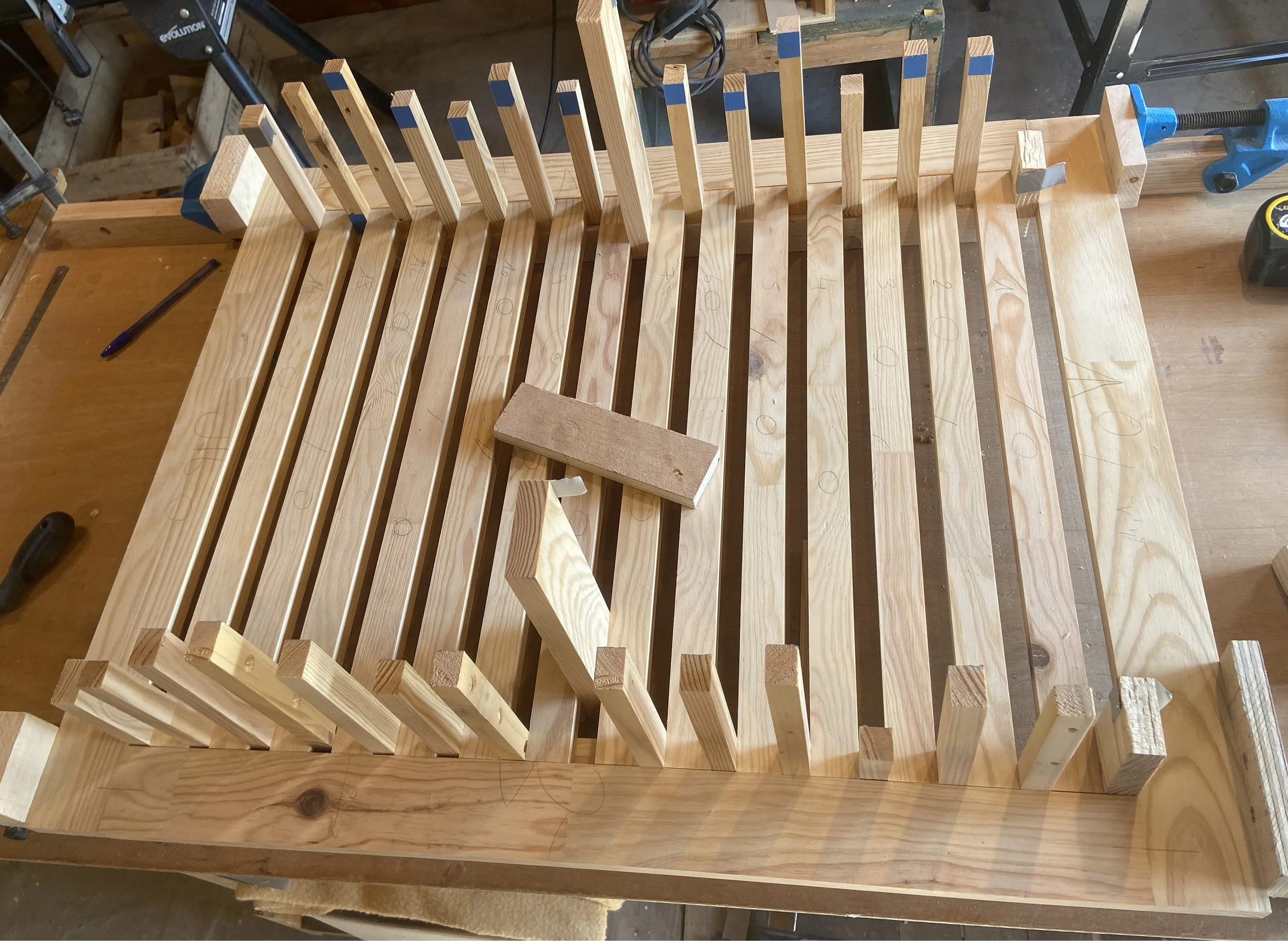Wooden slats and vertical support pieces clamped together on a workbench, with a small sanding block and pencil nearby, in a woodworking shop.