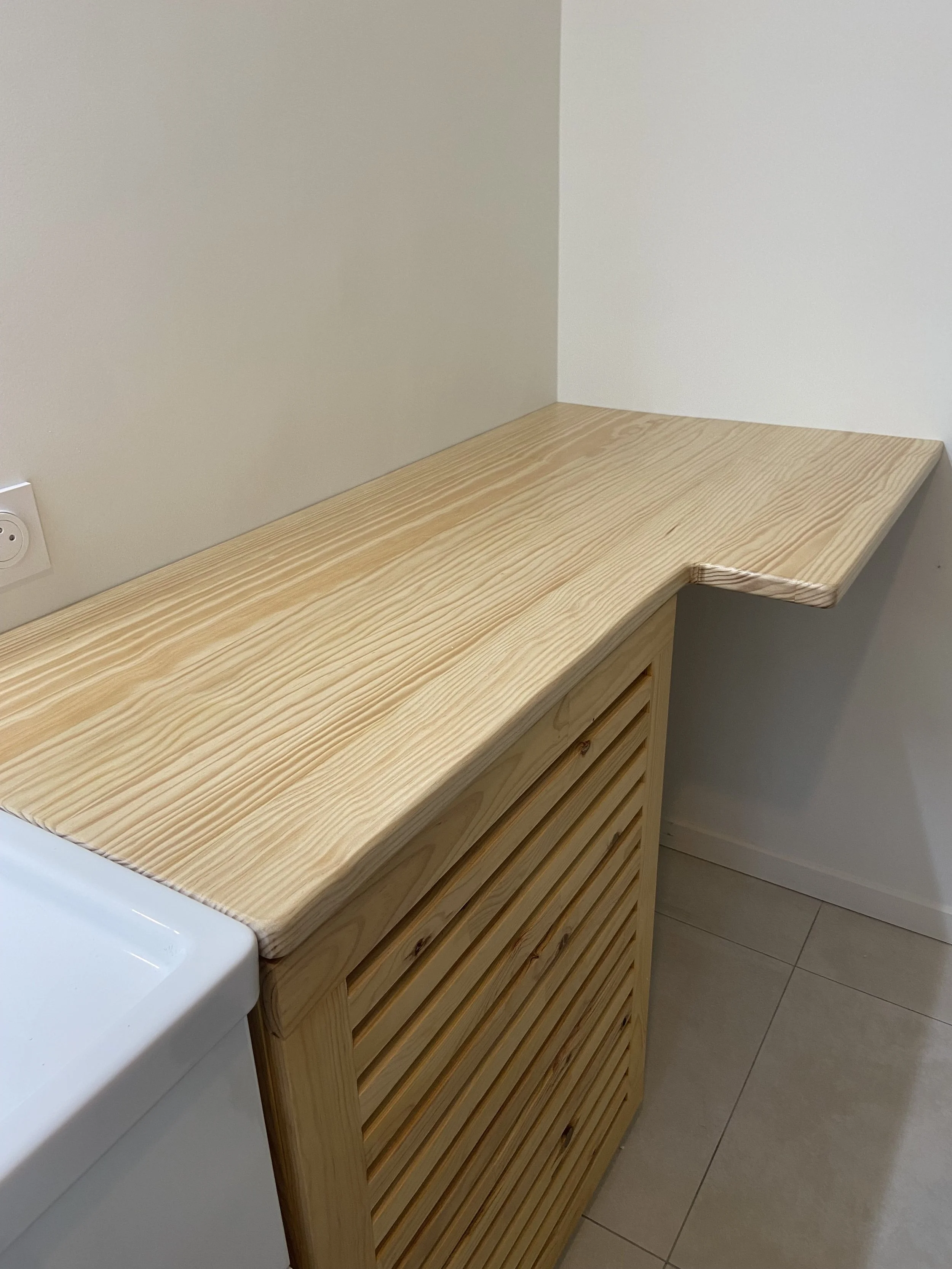 Wooden countertop with a rounded edge, adjacent to a white wall and tiled floor. Electrical outlet visible on the wall.