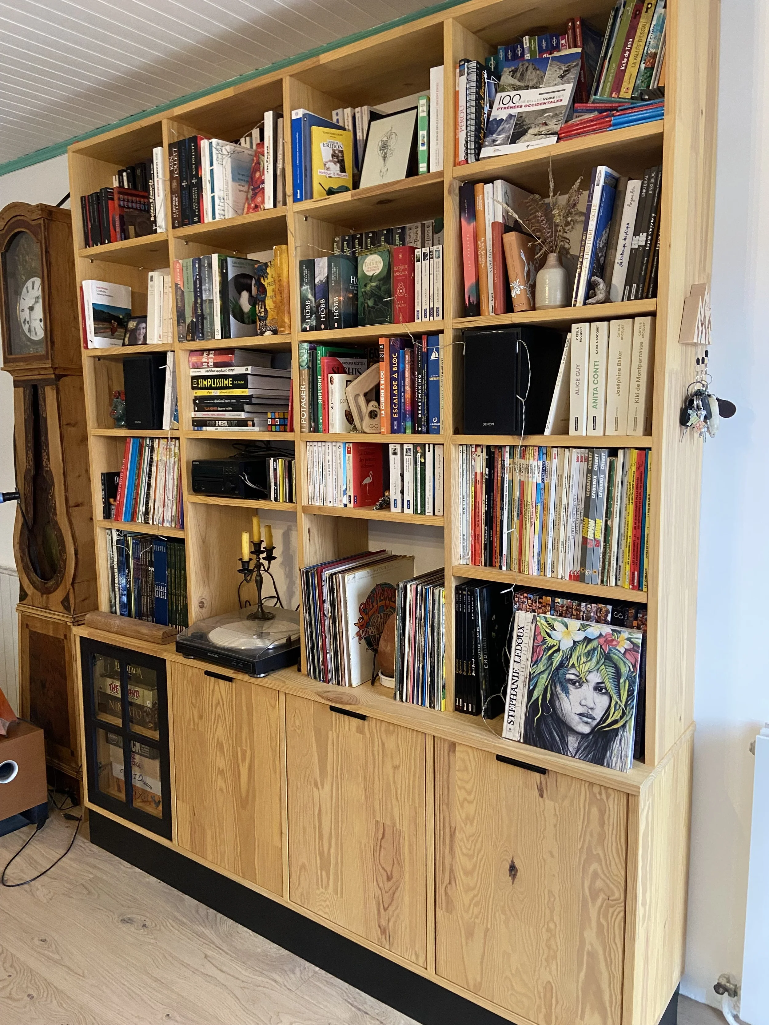 A wooden bookshelf filled with books, magazines, and decorative items including a vinyl record, a turntable, a candelabra, and a framed art piece, situated in a cozy room with a wooden floor and a vintage clock nearby.