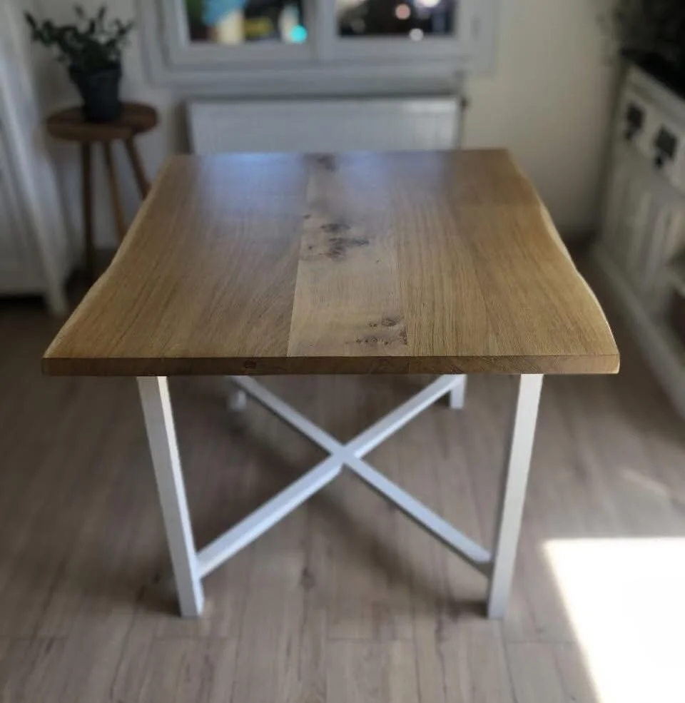 A wooden table with a white metal X-shaped leg design in a kitchen. The tabletop is made of light-colored wood with natural grain and knots. In the background, there is a window, a small side table with a potted plant, and kitchen appliances.