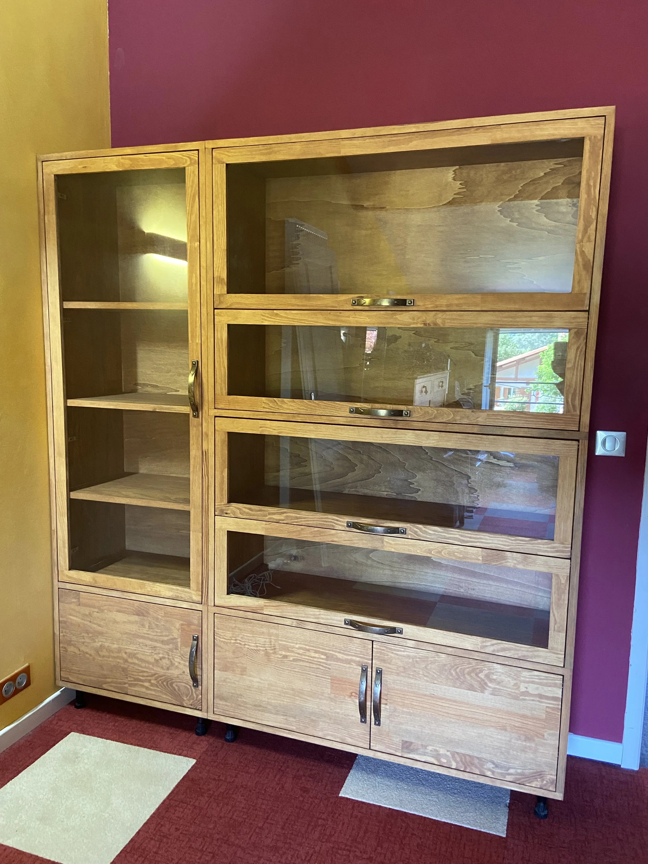 Empty wooden display cabinet with glass doors and shelves, positioned against a red and yellow wall on a red and beige carpet.