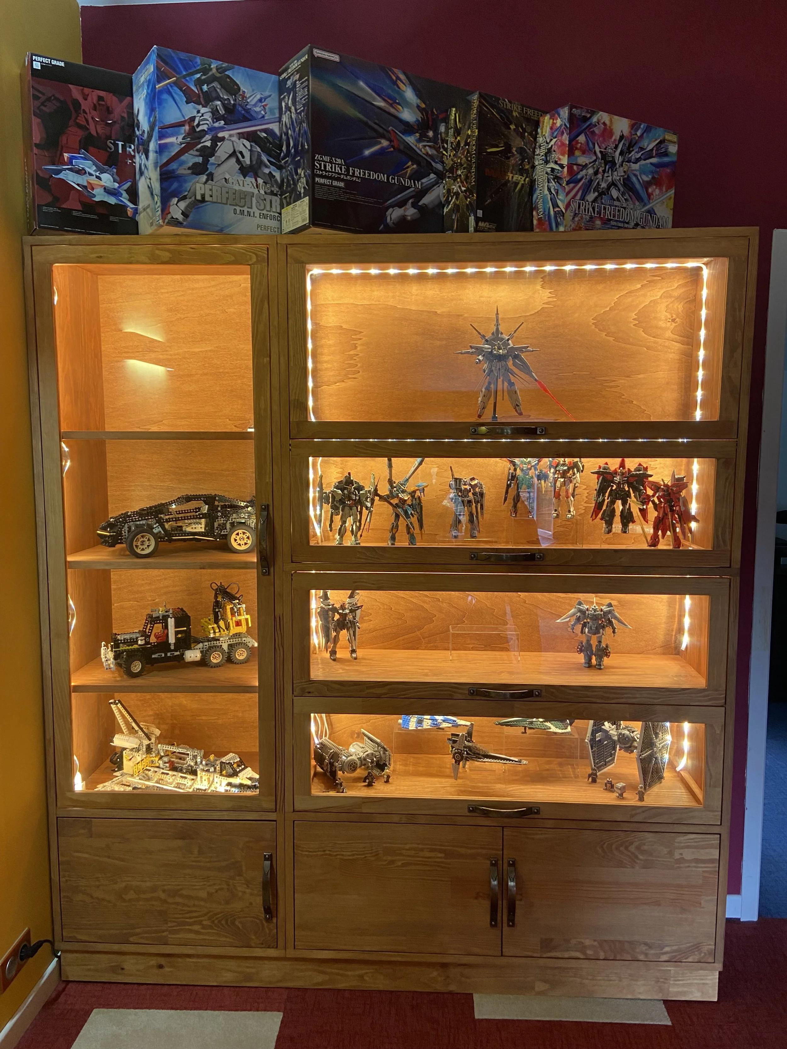 A wooden display cabinet with lights showcases various model robots and vehicles, with model boxes on top.