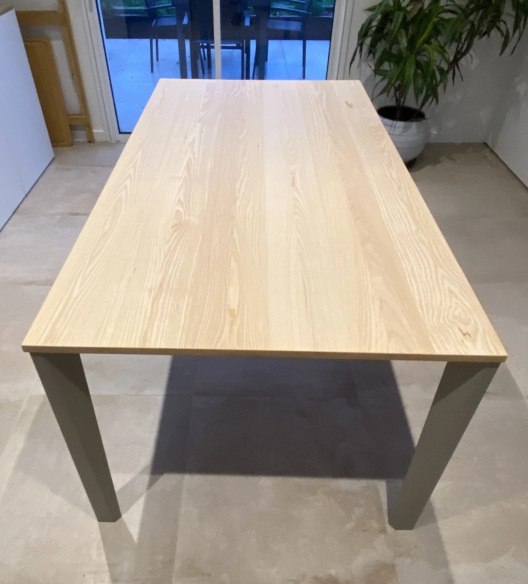 A wooden dining table with a light wood top and gray legs in a room with beige tile flooring, a sliding glass door in the background, and a potted plant on the right side.