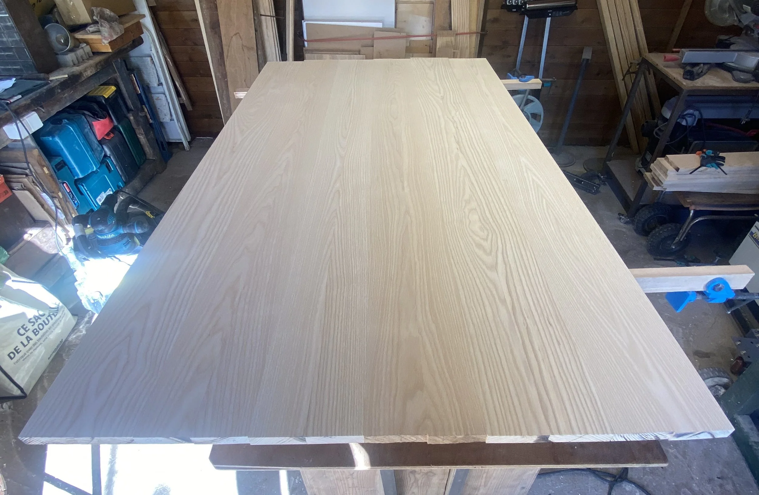 A large, light-colored wooden tabletop in a woodworking workshop, surrounded by various tools, equipment, and wood pieces.