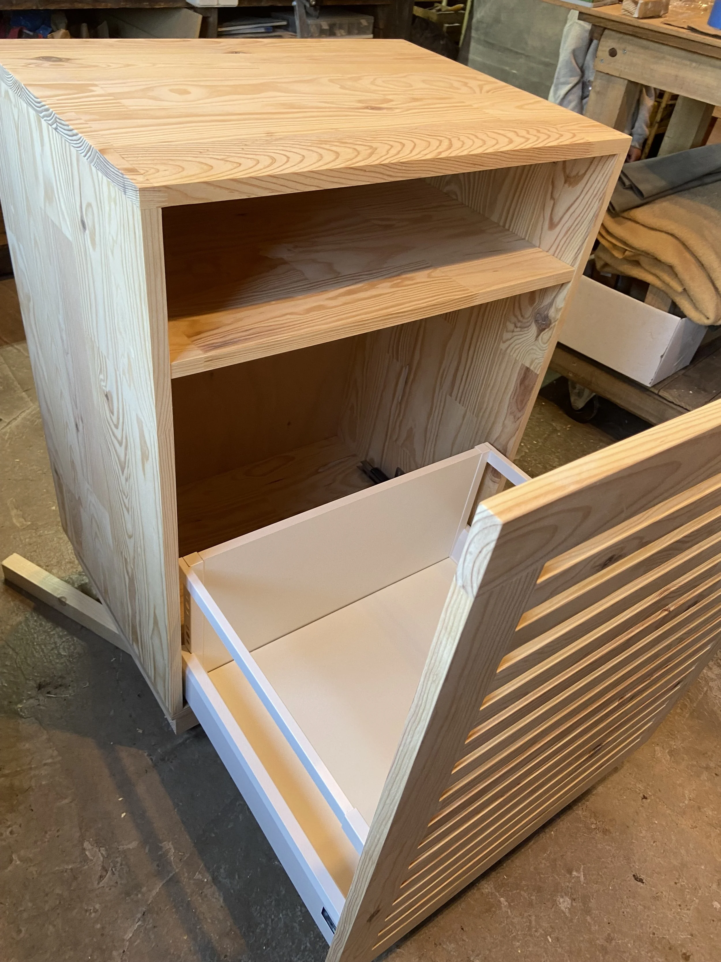 Unfinished wooden TV stand with open shelves and a drawer, positioned on a workshop floor.