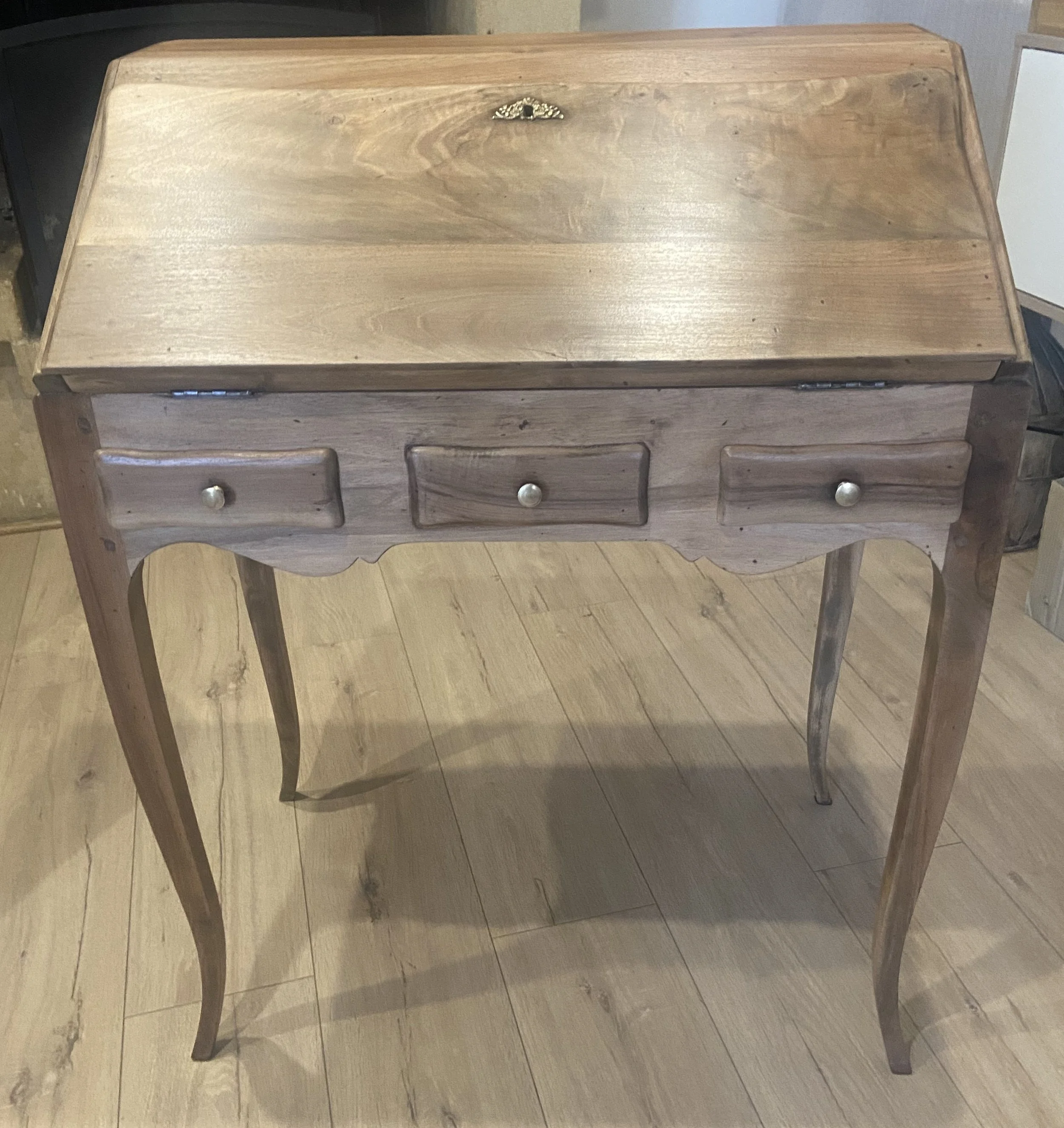 A wooden vintage writing desk with three small drawers and curved legs, situated on a wooden floor.