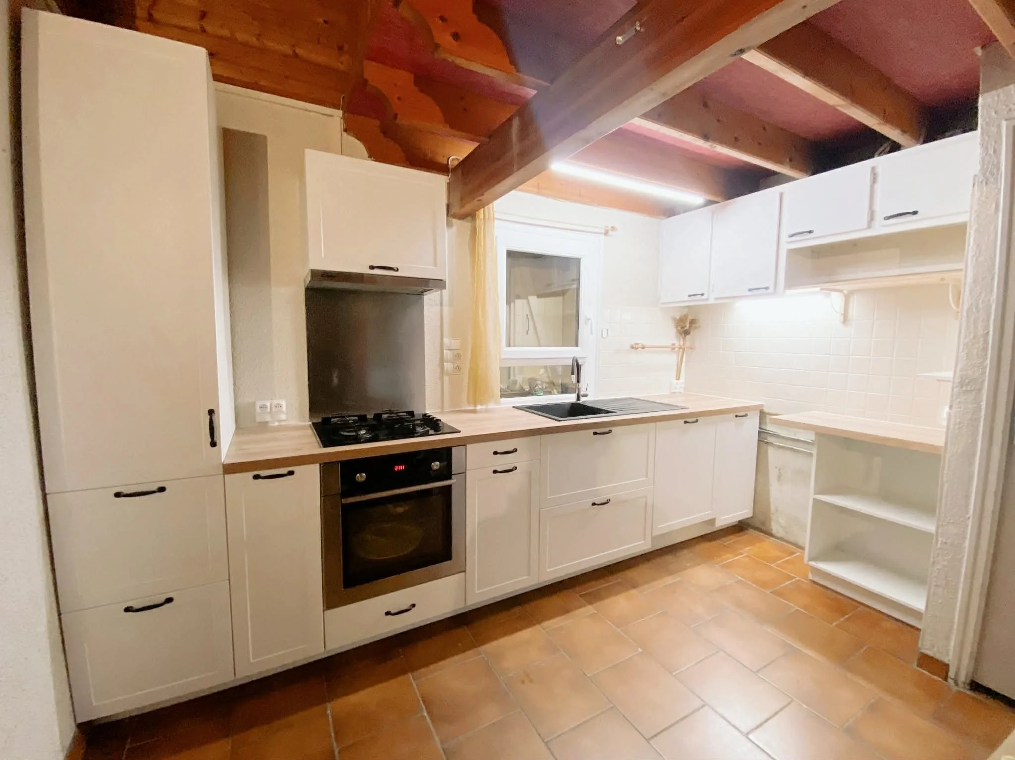 Empty kitchen with white cabinets, a wooden countertop, a black oven, gas stove, stainless steel sink, a window with a yellow curtain, wooden ceiling with exposed beams, and terracotta tiled floor.