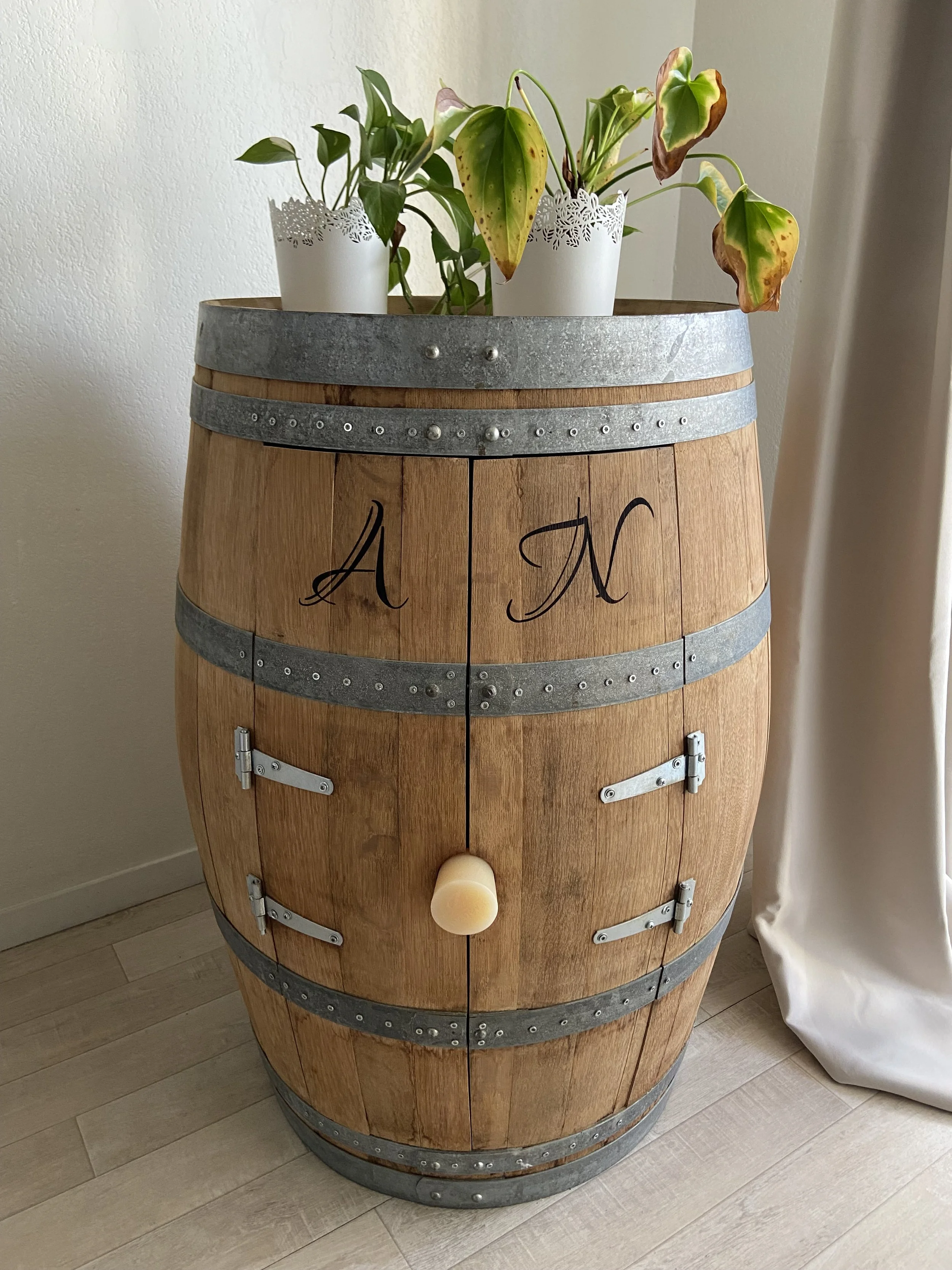 A wooden wine barrel with metal bands, used as a decorative table, with two potted plants on top. The barrel has the letters 'A N' written on it and a small round knob protruding from its front.