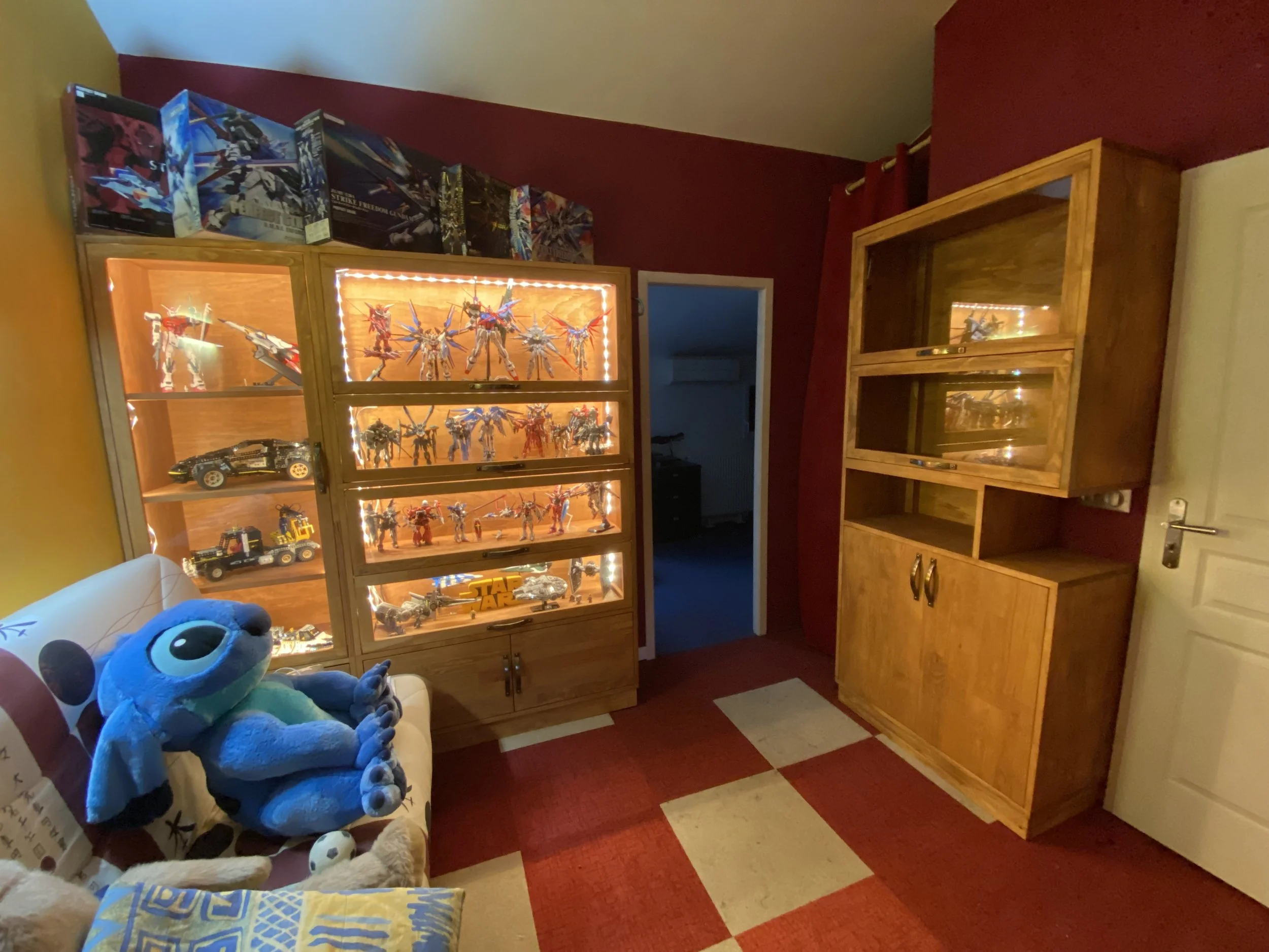 A room with red, yellow, and beige walls, featuring glass display cabinets filled with action figures and toys. There is a stuffed blue plush toy on a quilted chair or sofa near the foreground.