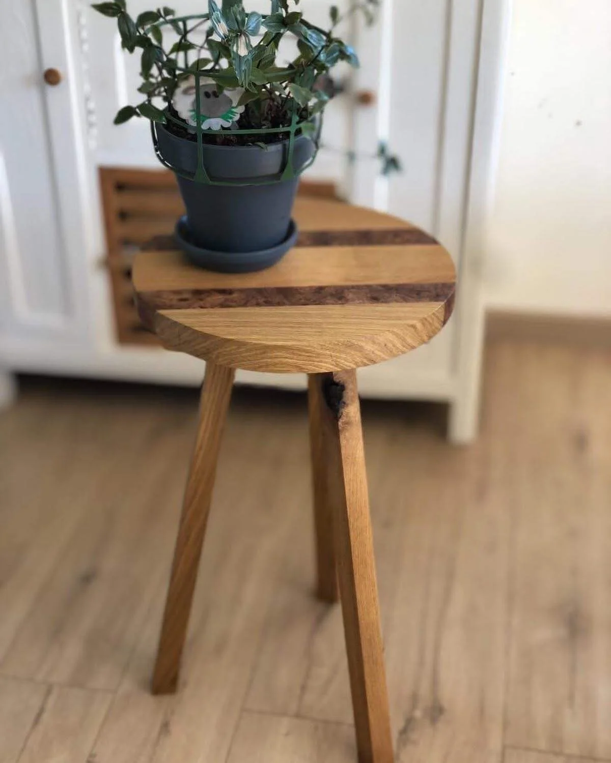 A small wooden tripod side table with a round top, holding a potted plant in a dark green container. The table is placed on a wooden floor against a light-colored background.