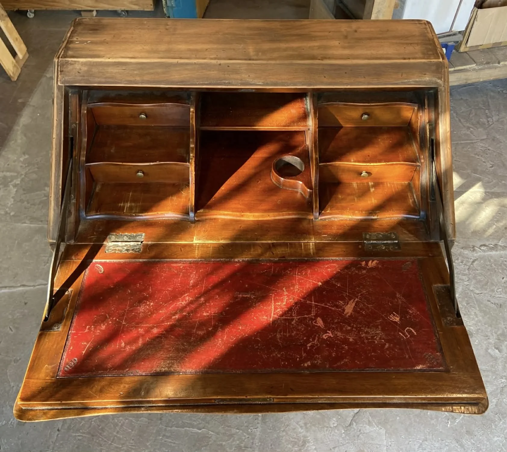 An antique wooden writing desk with multiple small drawers and a central circular hole, featuring a fold-down writing surface with a red leather inlay, situated on a concrete floor.
