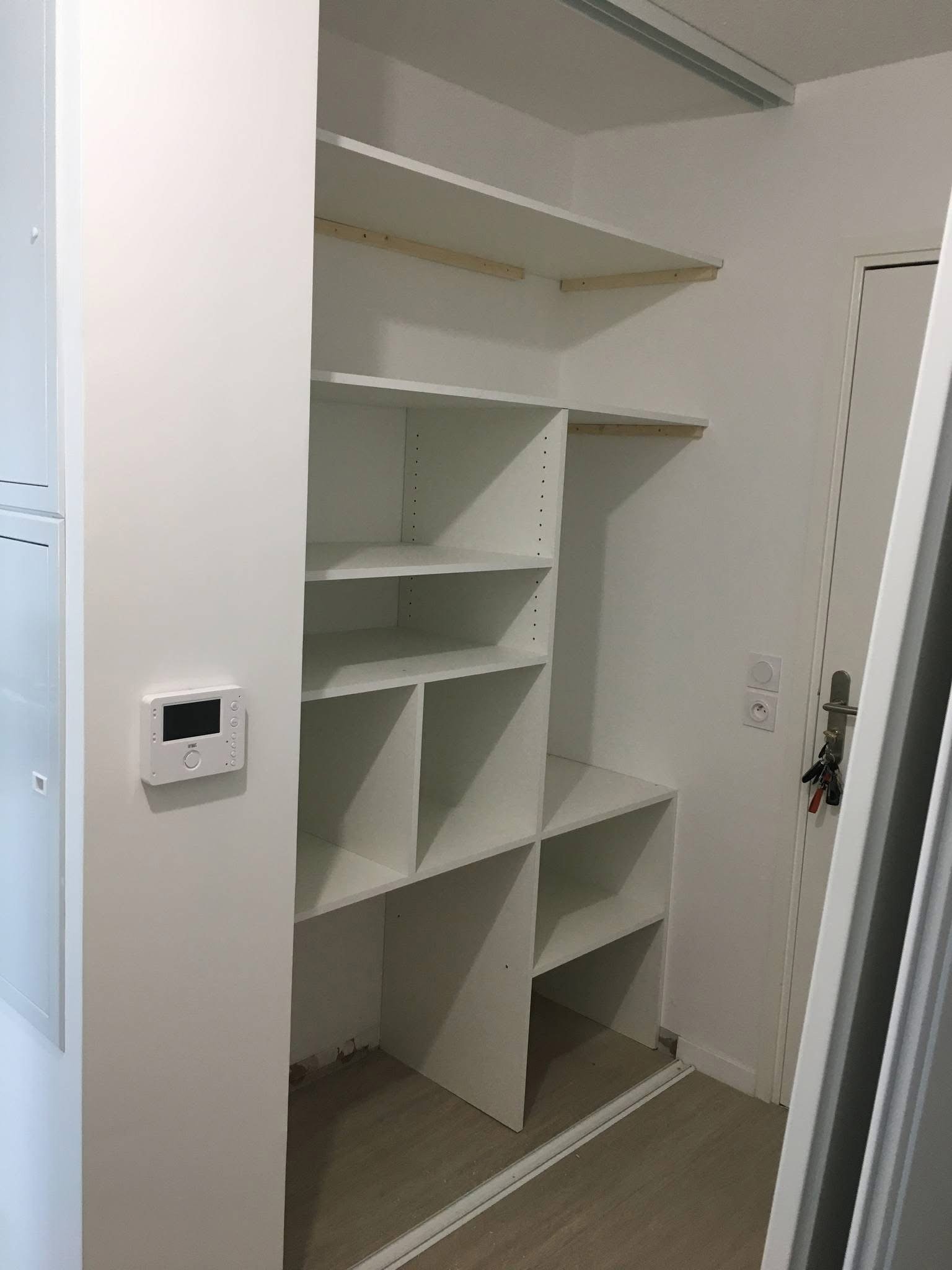 Empty white shelving unit with multiple compartments located next to a white door and a control panel on the wall.