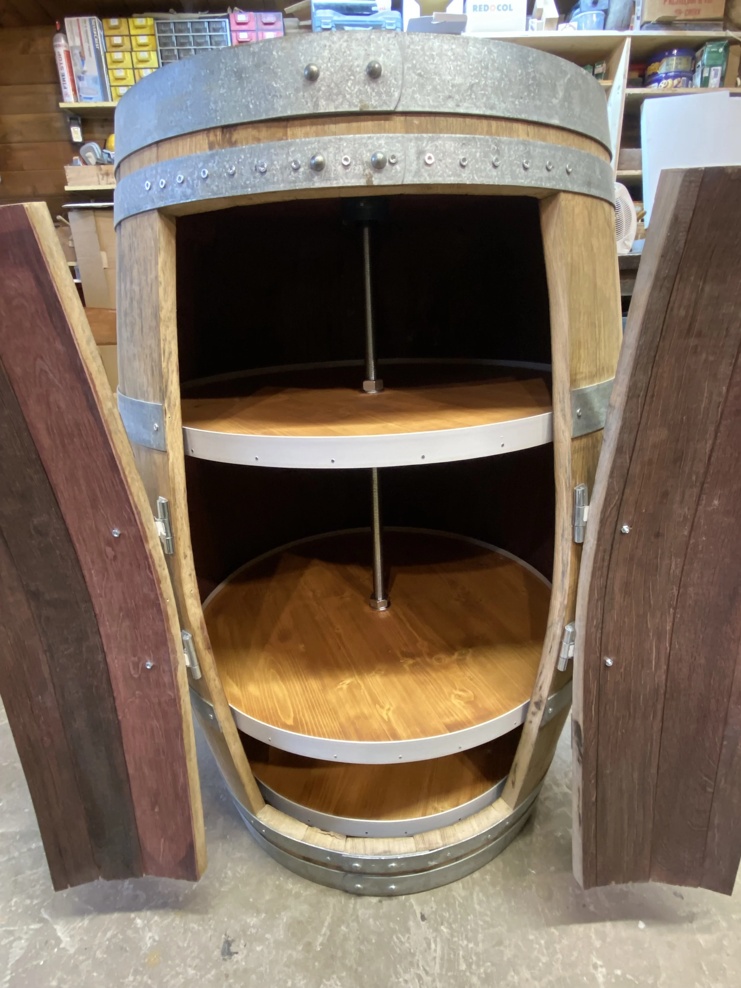 A custom wine barrel cabinet with open wooden doors revealing two circular shelves inside, with metal supports and hinges, in a store or workshop setting.