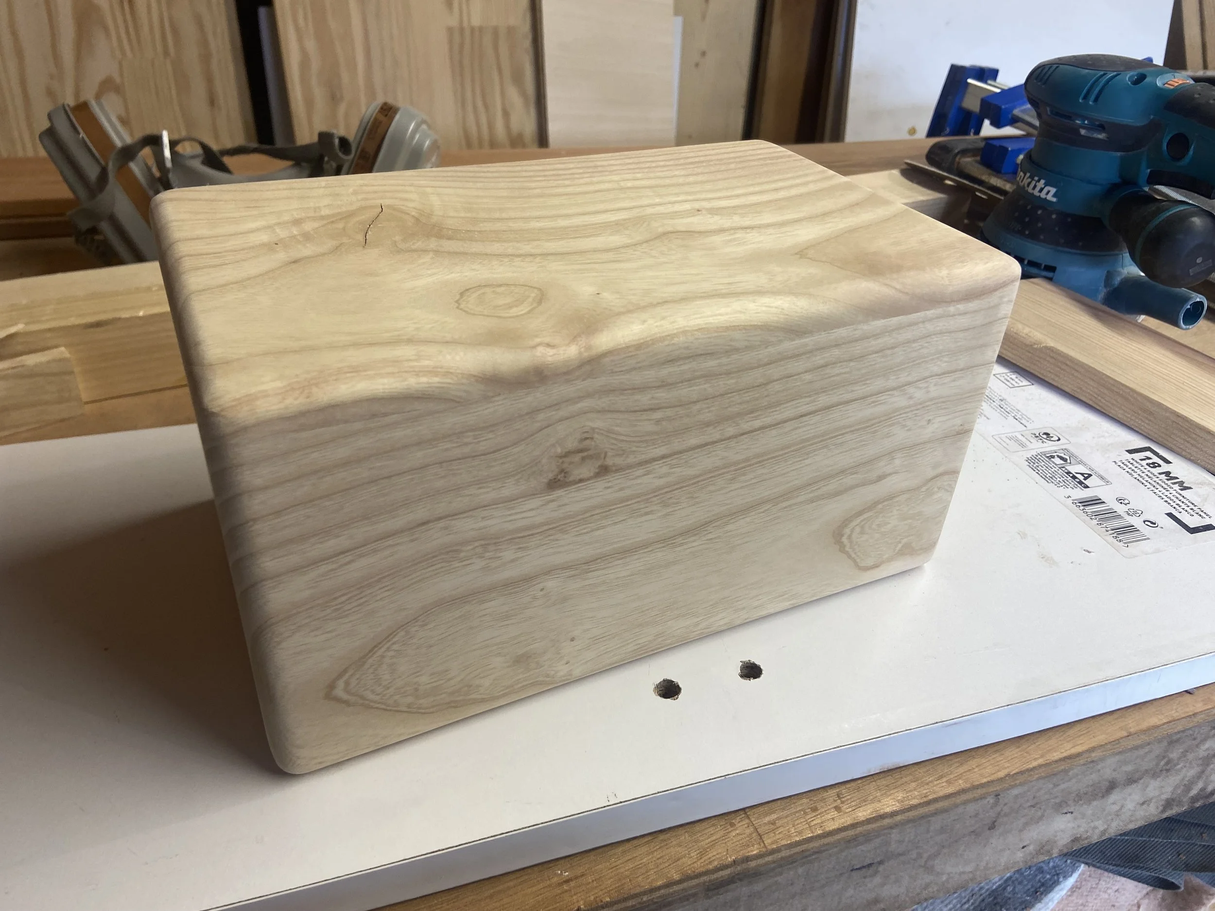 A rectangular block of unfinished light-colored wood on a workbench in a woodworking shop, with power tools and wood pieces in the background.