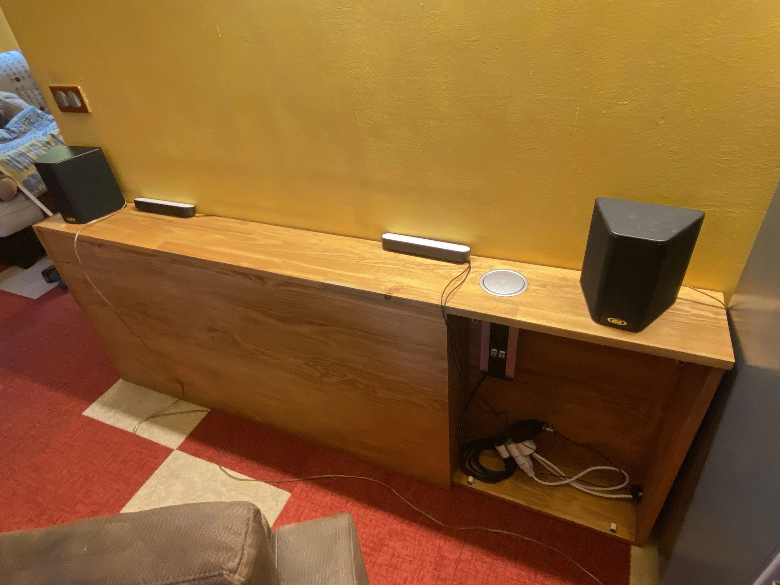 Wooden sideboard with black speakers, a small white device, and a circular metallic object on top, with electronic cables hanging underneath. A yellow textured wall is behind it, and part of a brown sofa and a red patterned carpet are visible.