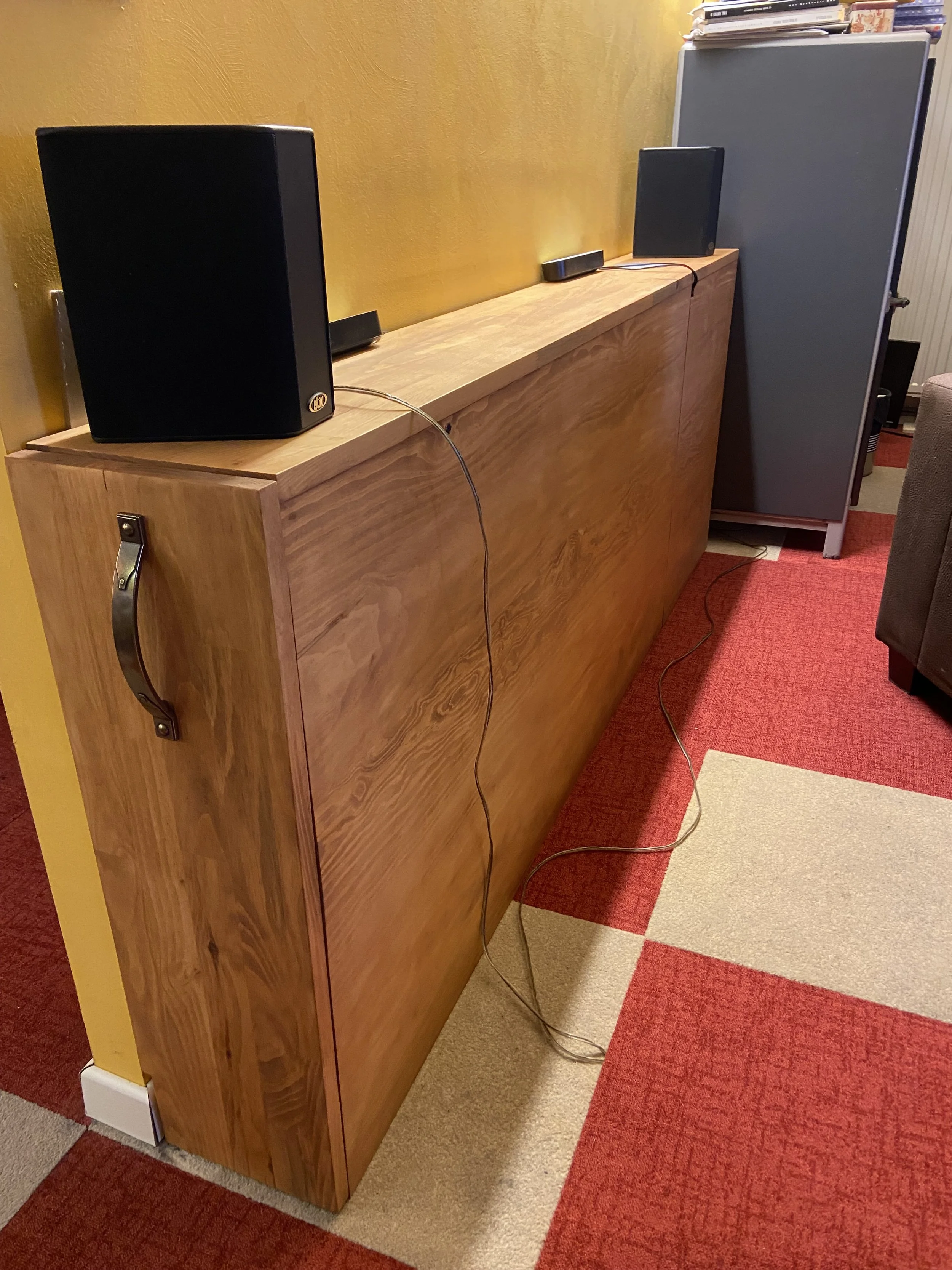 A wooden storage cabinet with a handle on the side, two black speakers on top, and a remote control between them. A yellow wall behind and a red, beige, and gray checkered carpet on the floor.
