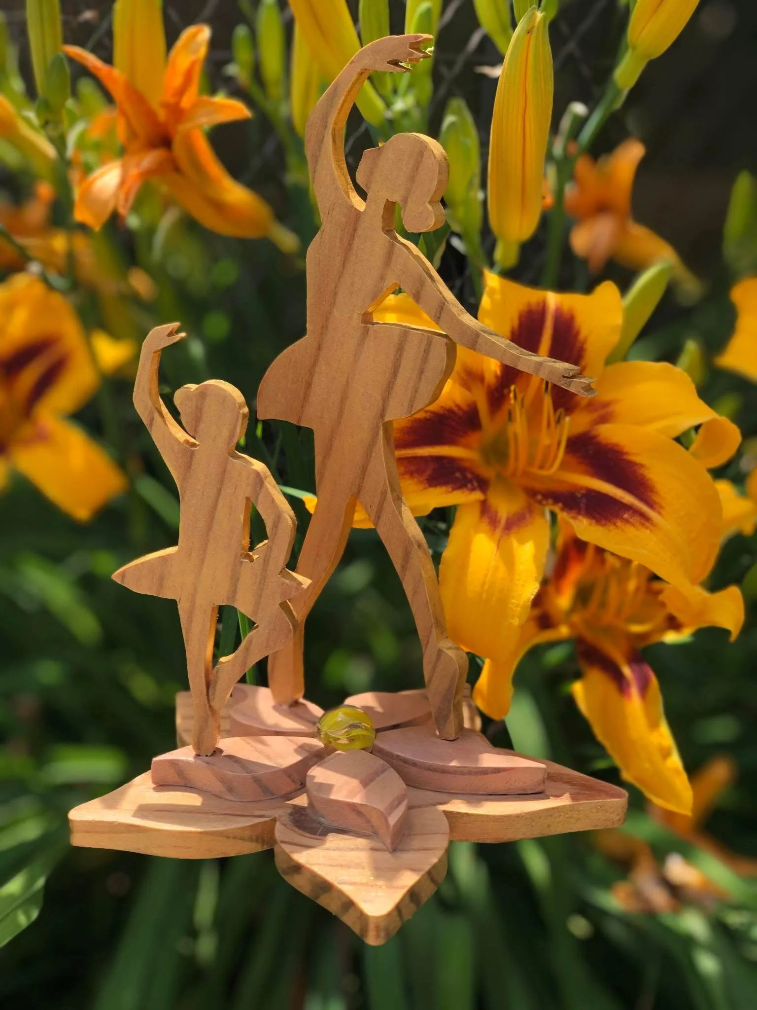 Wooden sculpture of a ballerina and a girl, positioned among yellow flowers with maroon accents.