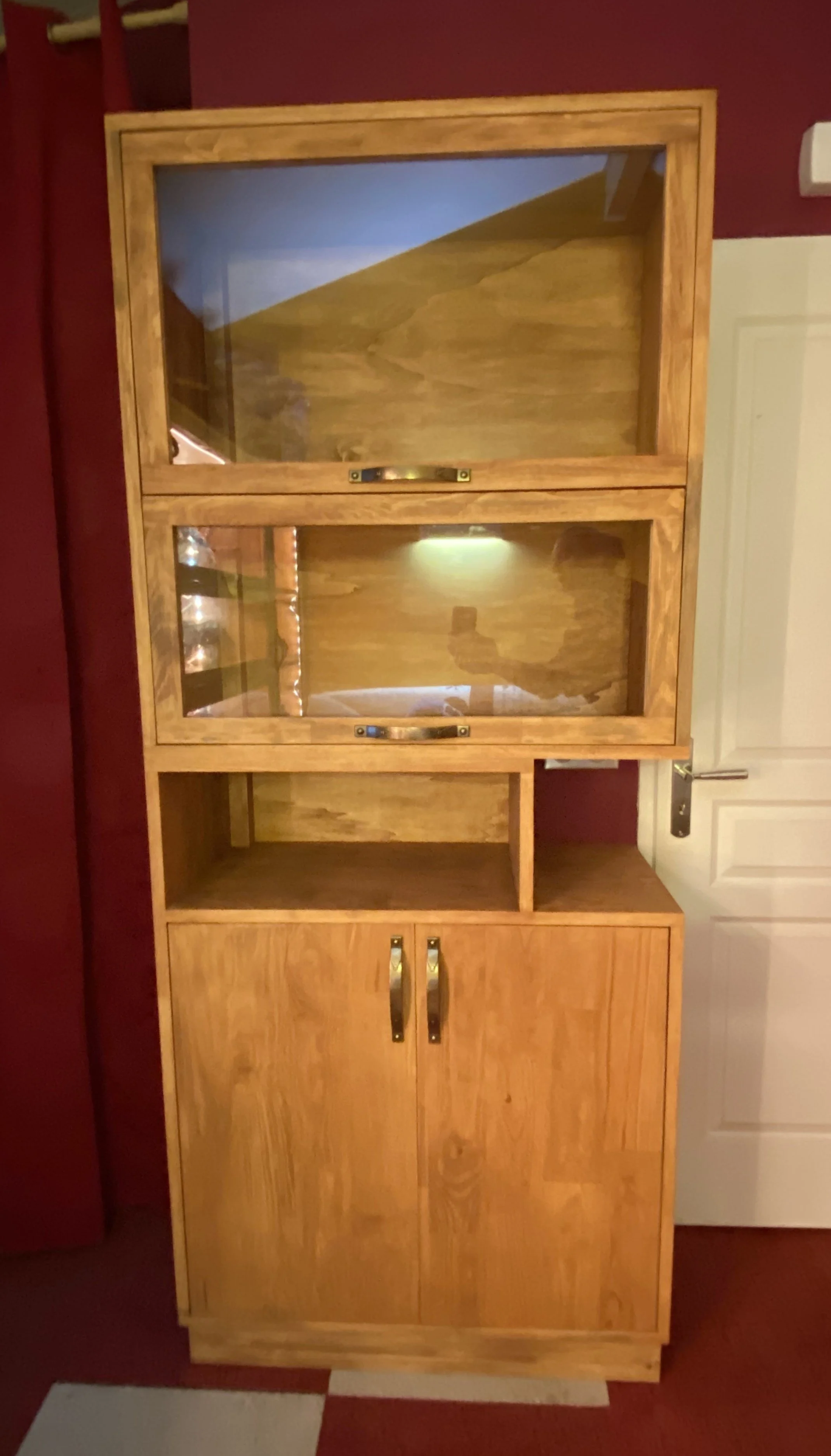 A wooden cabinet with glass doors on the top section and wooden cabinet doors below, set against a red wall.