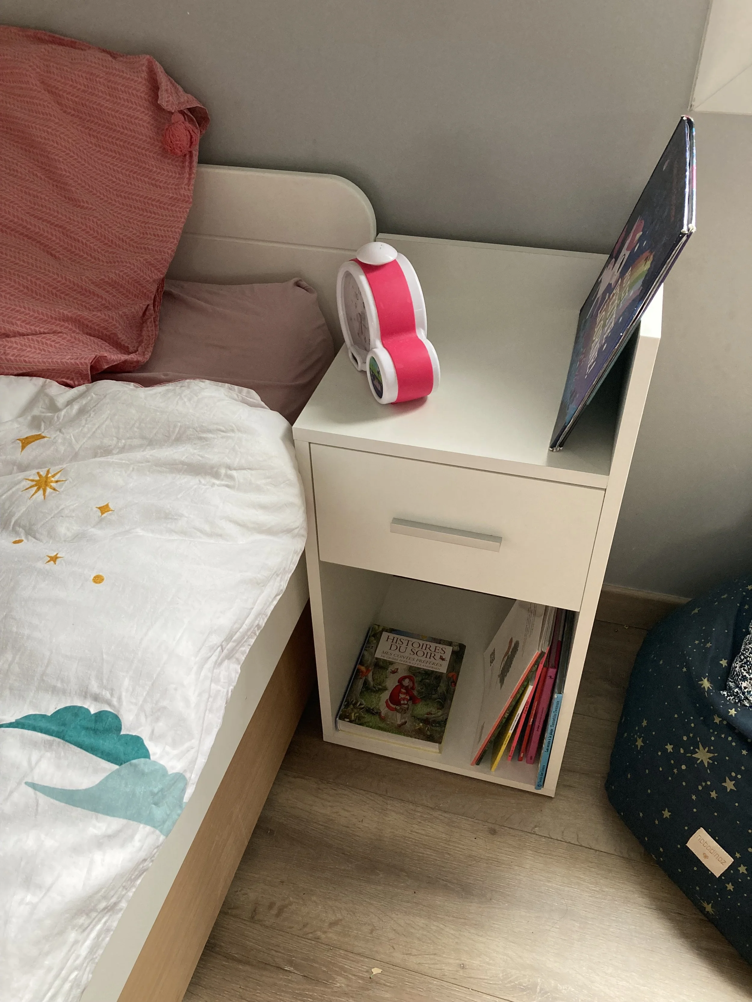 A white bedside table next to a bed with pink and white bedding, with a pink and white alarm clock and a tablet on top. The lower shelf holds books and magazines, including one titled 'Histoires du Soir.' Part of a blue star-patterned bean bag is vis