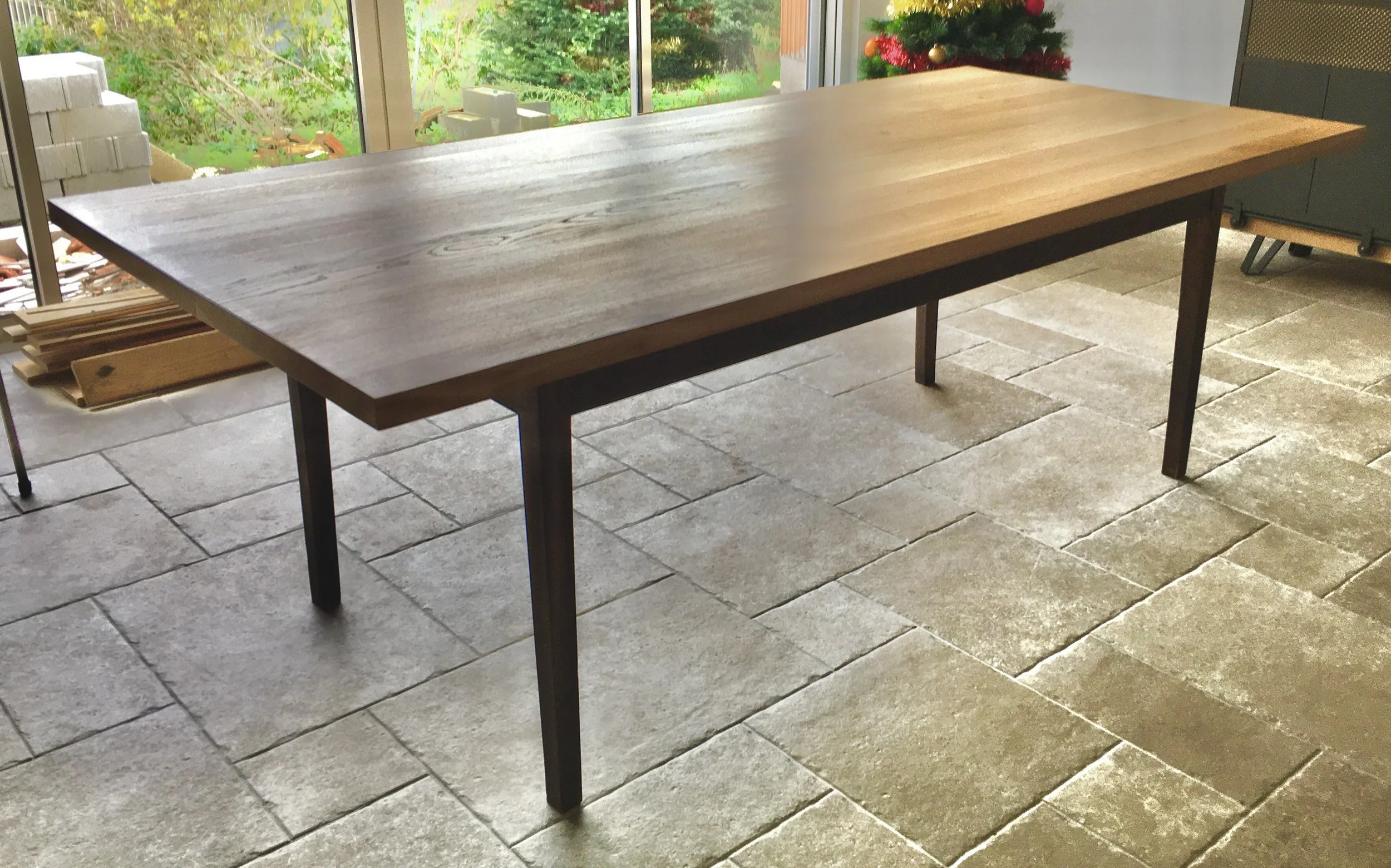 A large wooden dining table with a natural finish in a room with stone tile flooring and a Christmas tree in the background.