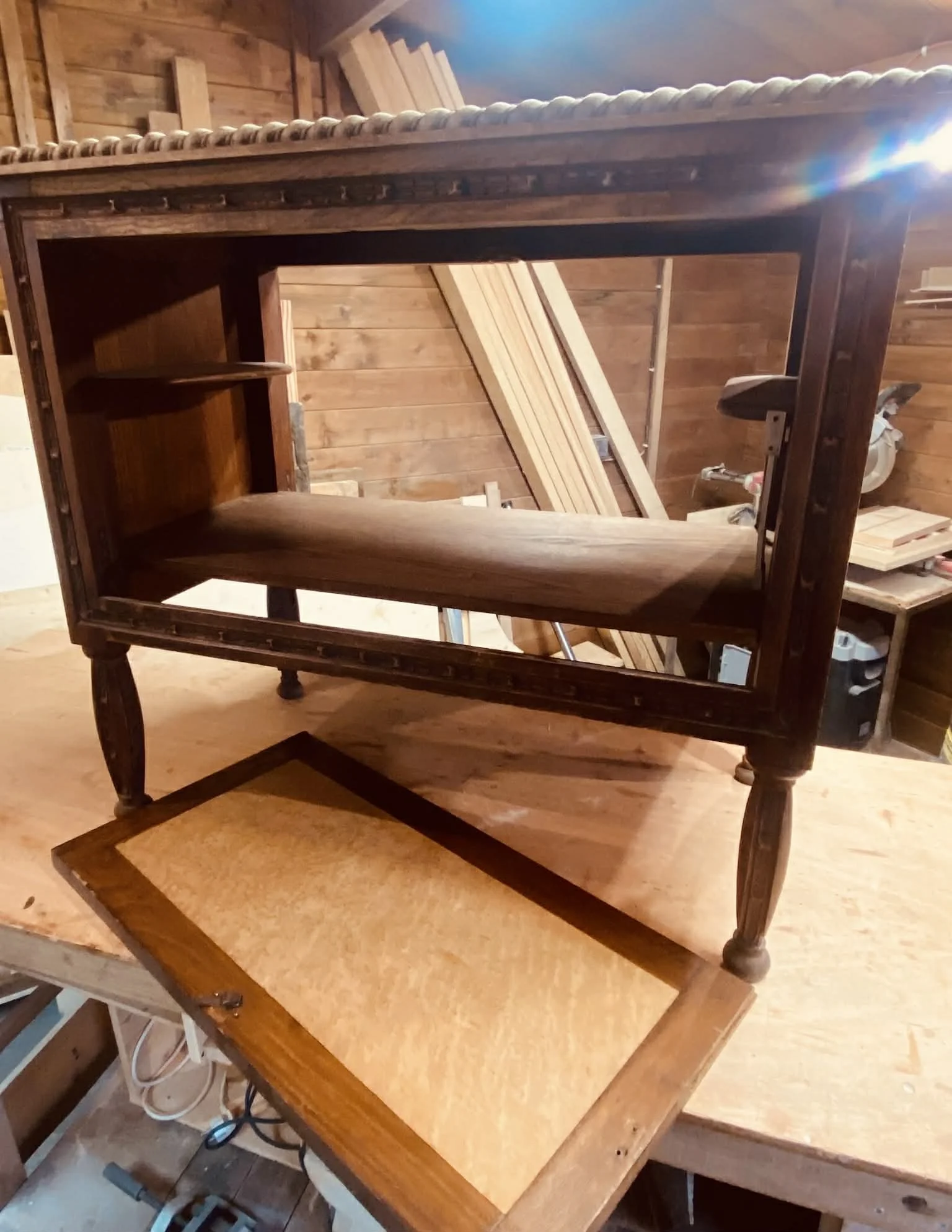 A wooden table with a darker finish sits on a workbench in a woodworking shop, with a wooden frame or drawer on top. The background shows wooden planks and tools in a workshop setting.
