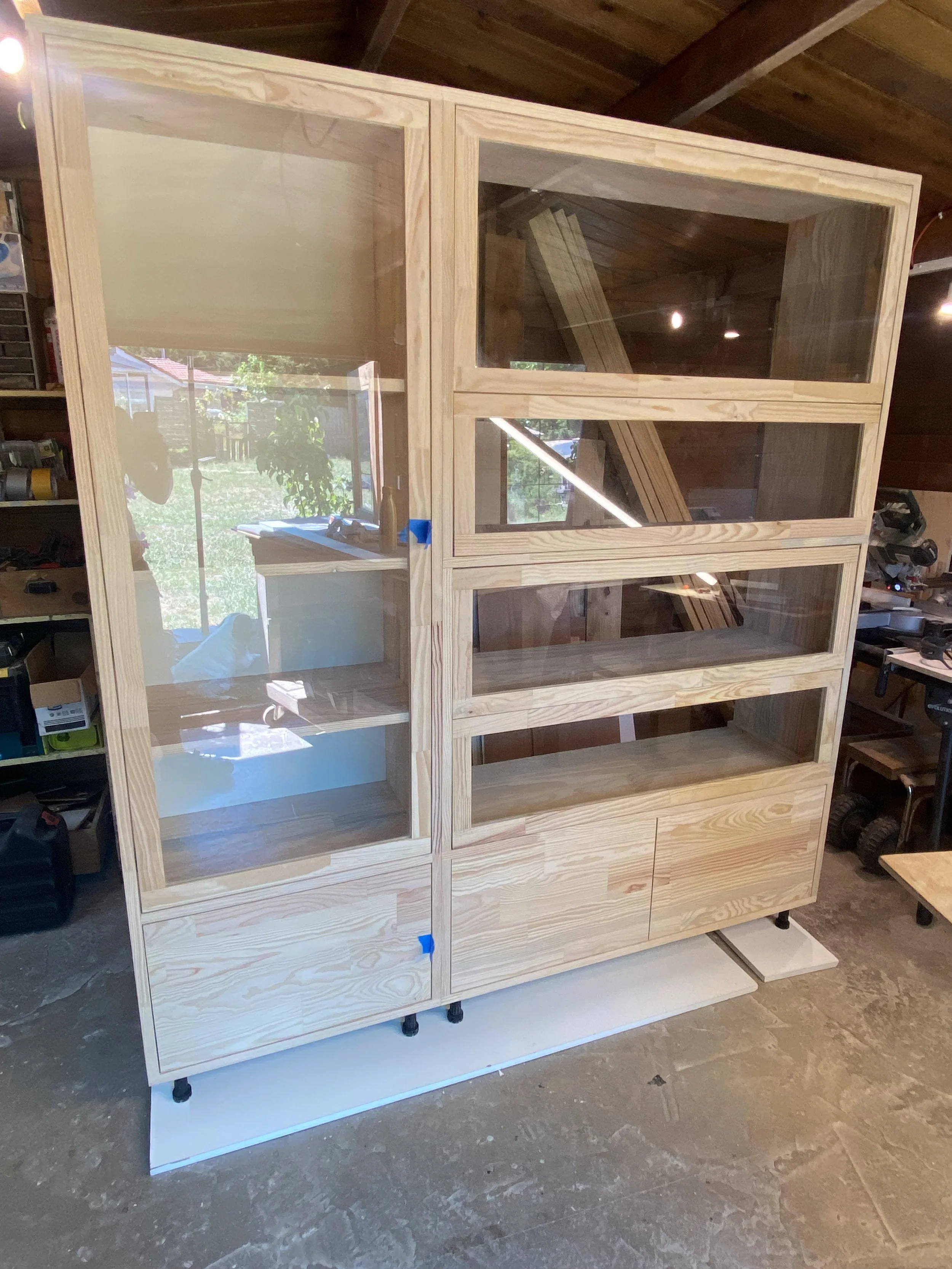unfinished wooden bookshelf with glass doors and open shelves, placed on a white base in a workshop.