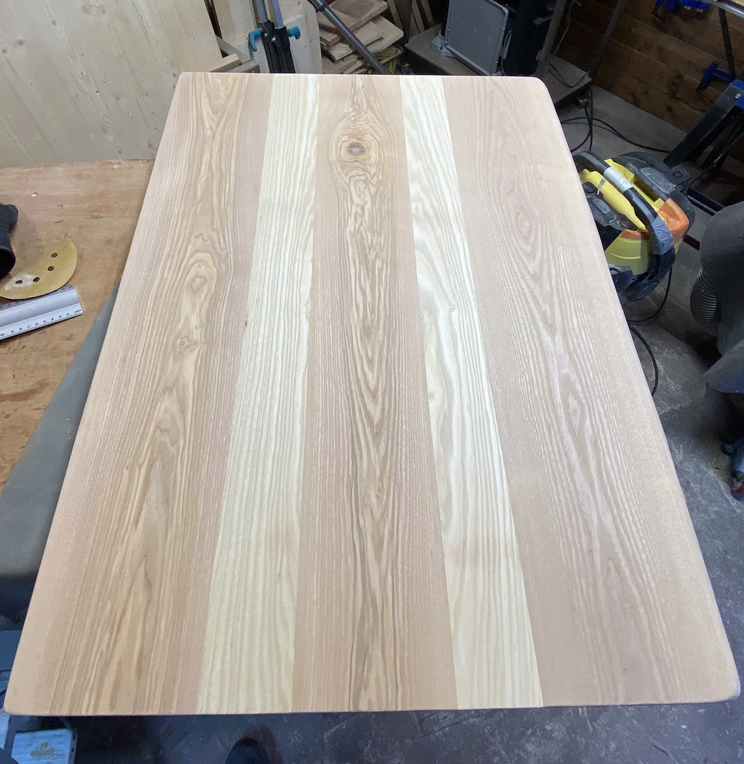 A light-colored wooden table with visible natural wood grain, sitting in a workshop or woodworking space.