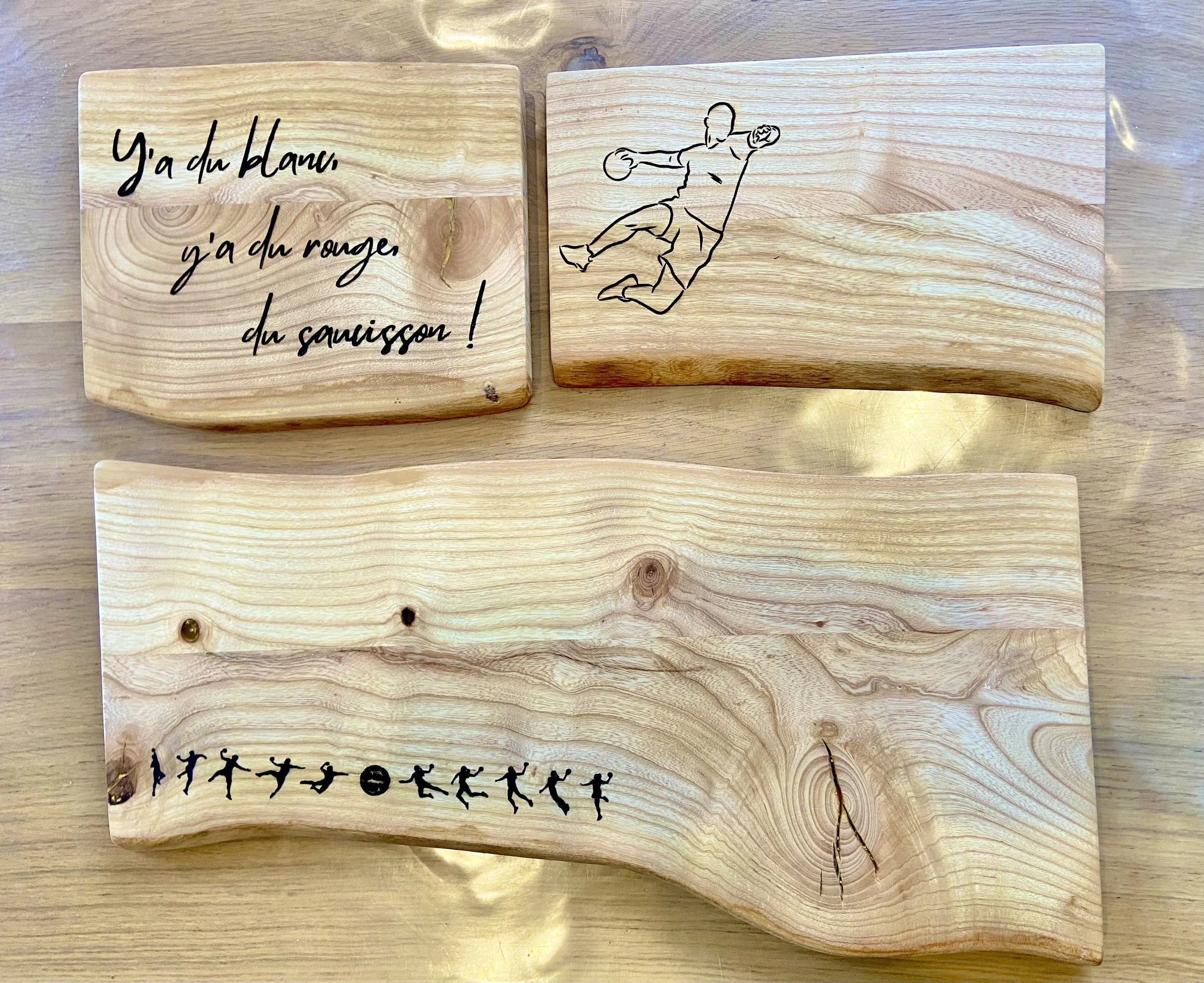 Three wooden plaques with engraved and painted designs on a wooden surface. The top left plaque has French text that reads 'Y'a du , y'a du rouge, du saucisson!' in black cursive. The top right plaque depicts a line drawing of a person diving or jump