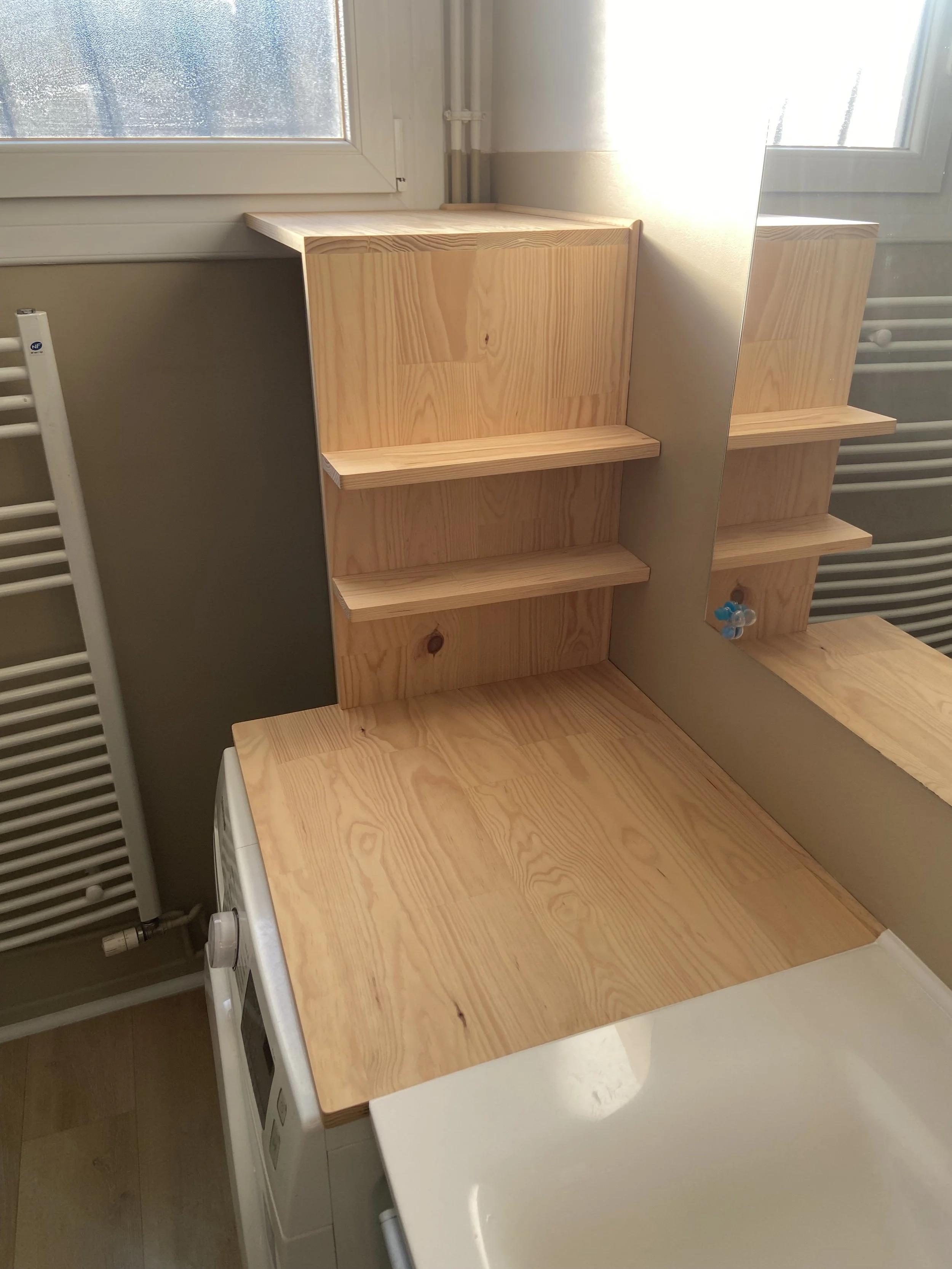 Wooden countertop with built-in shelving unit behind a washing machine, next to a mirror and a window in a laundry room.