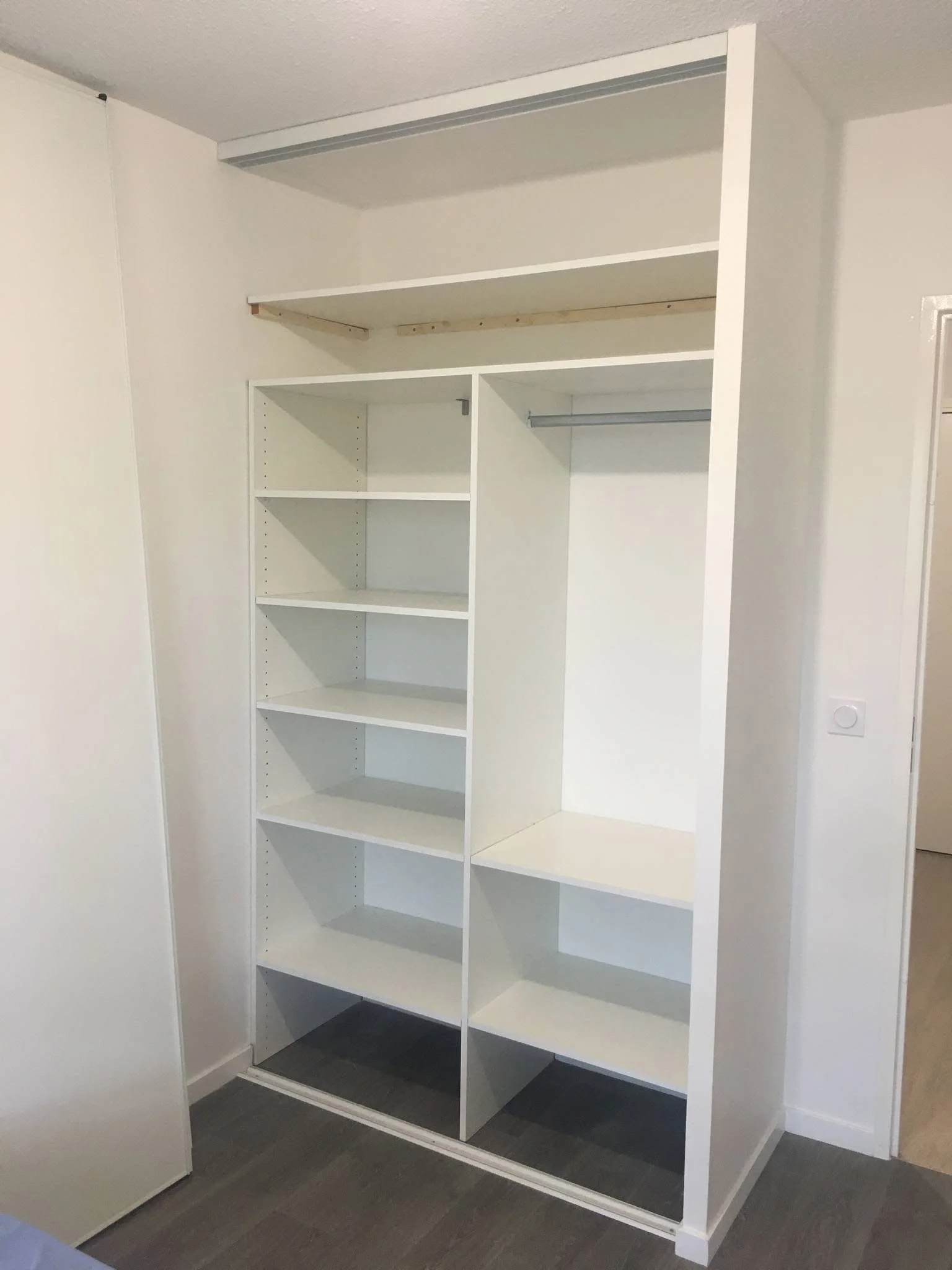 Empty white built-in closet with shelves and hanging space, set against white walls and dark flooring.