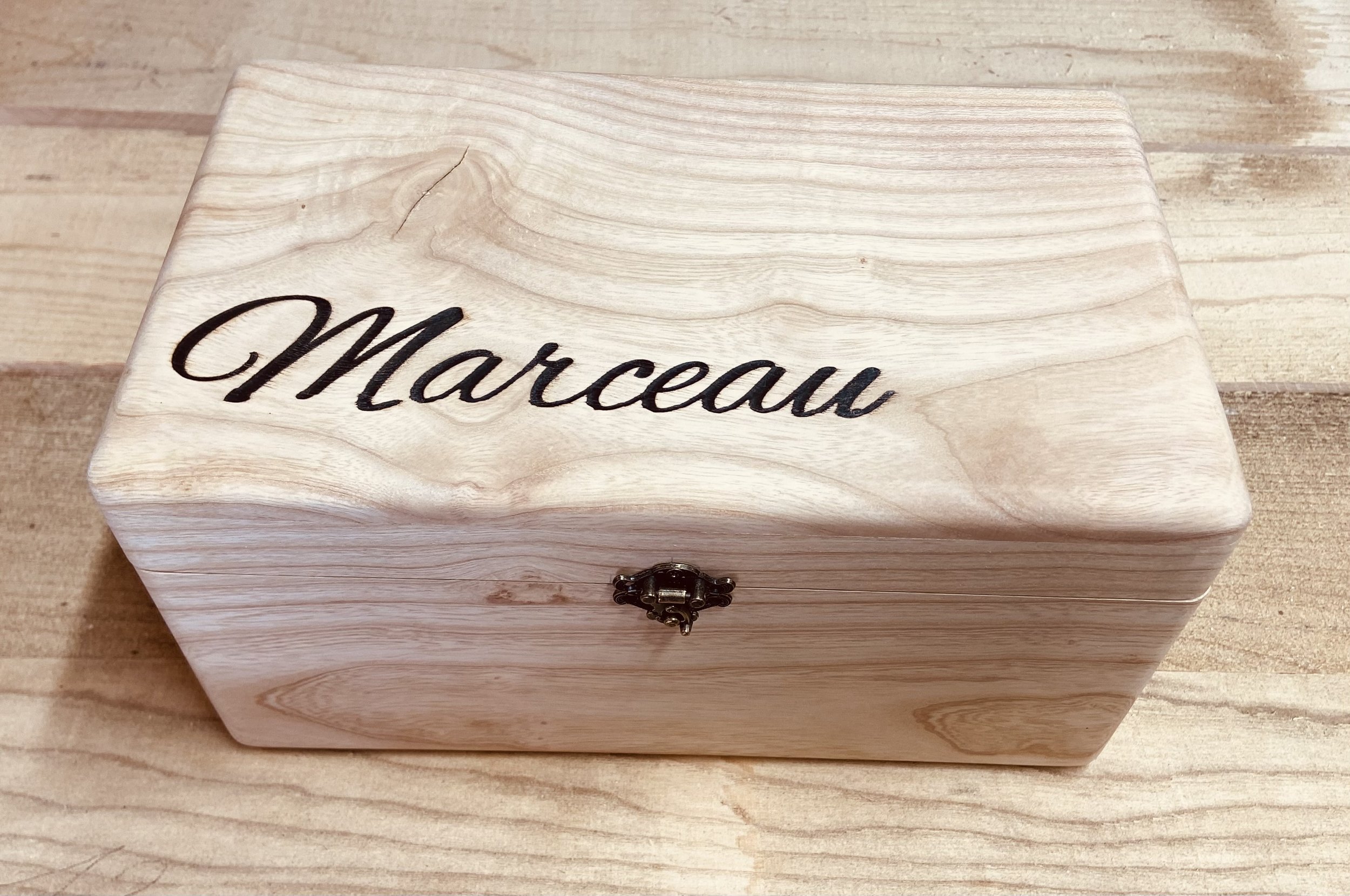 A light-colored wooden jewelry box with a black latch, engraved with the name 'Marceau' in black cursive letters on the lid, placed on a wooden surface.