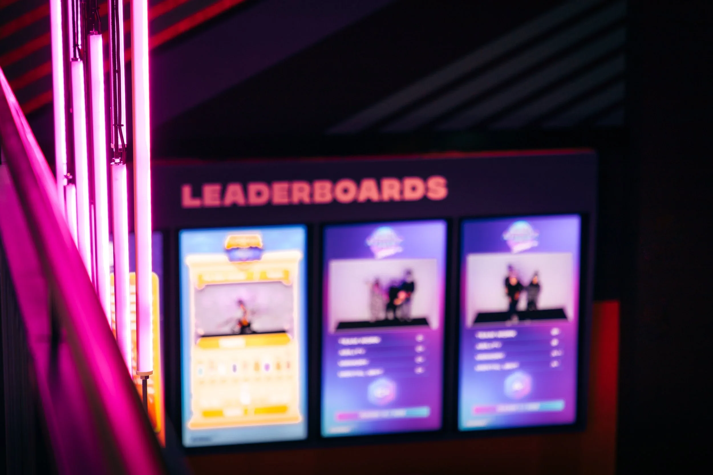 A gaming arcade leaderboard display with three screens showing players and game stats, illuminated by pink neon lights.